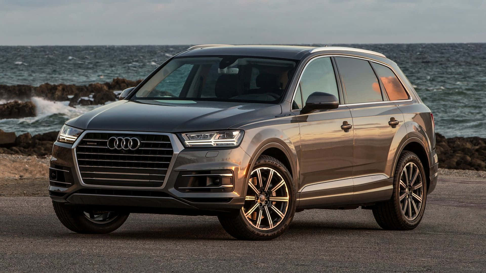 Audi Q7 桌布 壁紙 — 一輛時尚灰色奧迪Q7 SUV停泊在海岸公路上，背後有海浪拍打岩石。