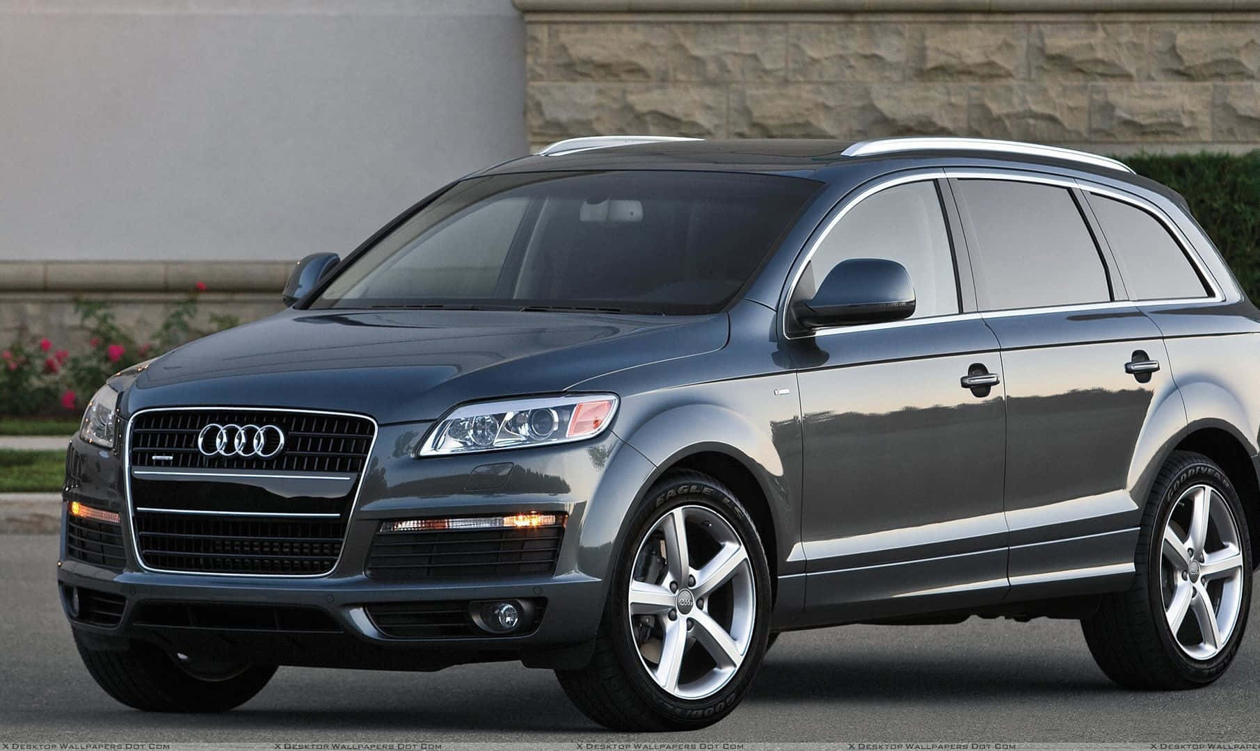 Audi Q7 桌布 壁紙 — 一輛時尚的灰色奧迪Q7 SUV停放在人行道上，背景有石牆和花朵。