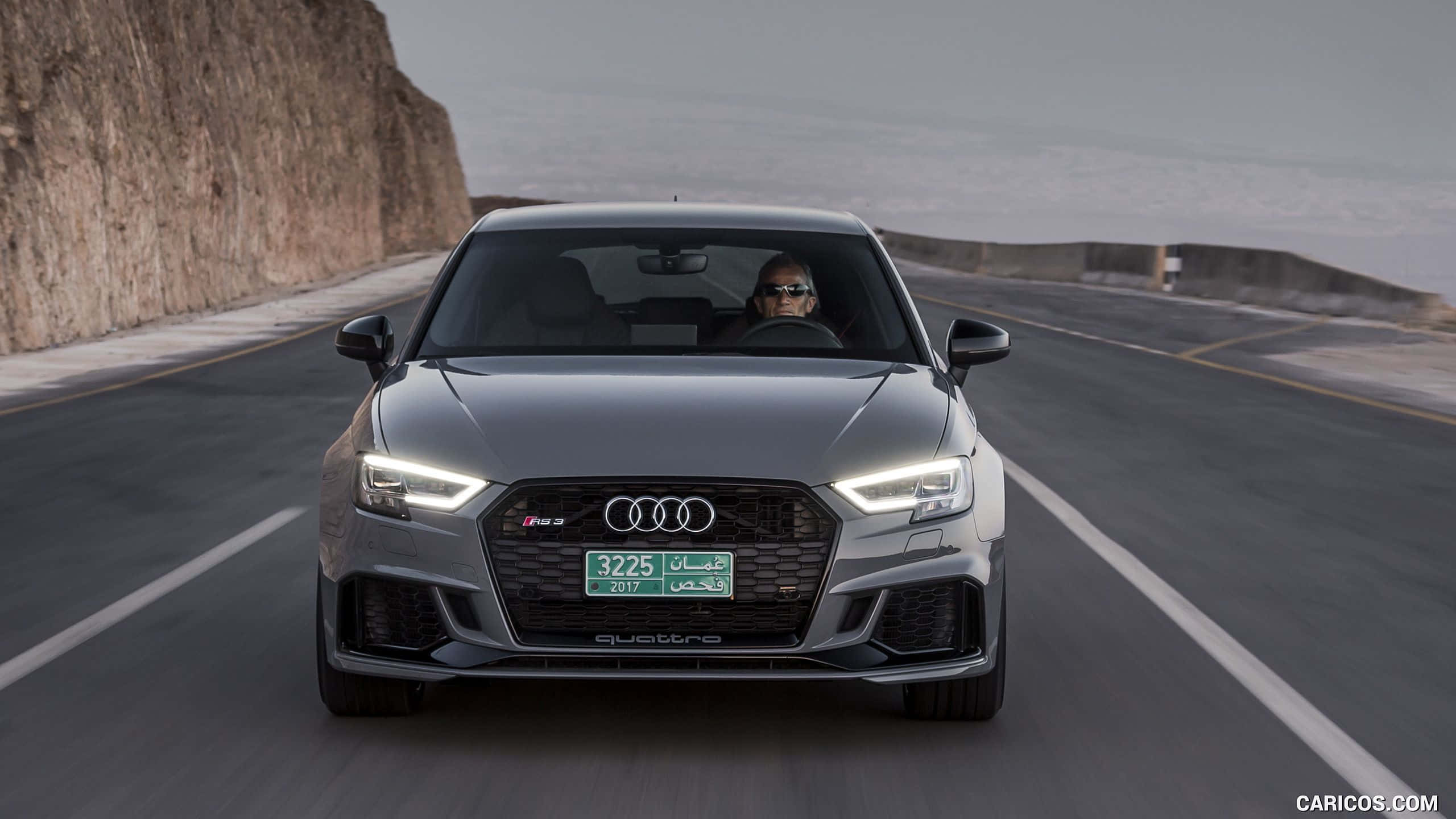 Audi Rs3 桌布 壁紙 — 一輛流線型灰色Audi RS3 quattro沿著海岸公路飛馳，一側是崎嶇懸崖，另一側是海洋。