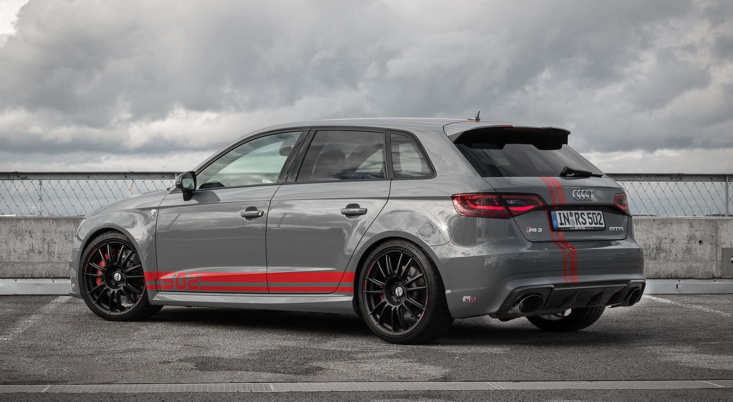 Audi S3 桌布 壁紙 — 一輛時尚的灰色奧迪RS3 Sportback，帶有醒目的紅色條紋，停在多雲天空下的混凝土屋頂上。