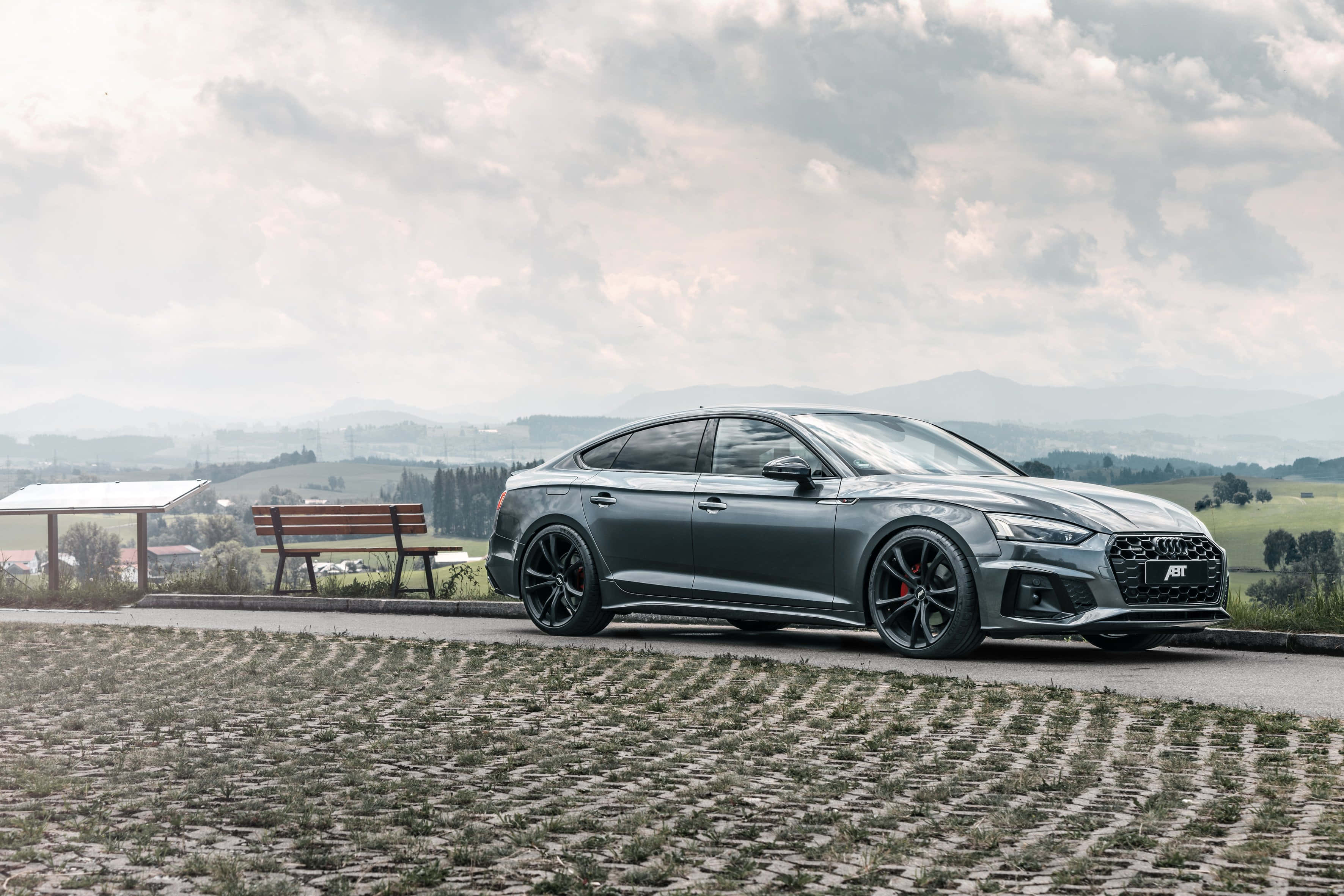 Audi A5 桌布 壁紙 — 一輛時尚的灰色奧迪轎車配黑色輪圈，停在鋪砌道路上，俯瞰風景如畫的鄉村山谷，天空多雲。