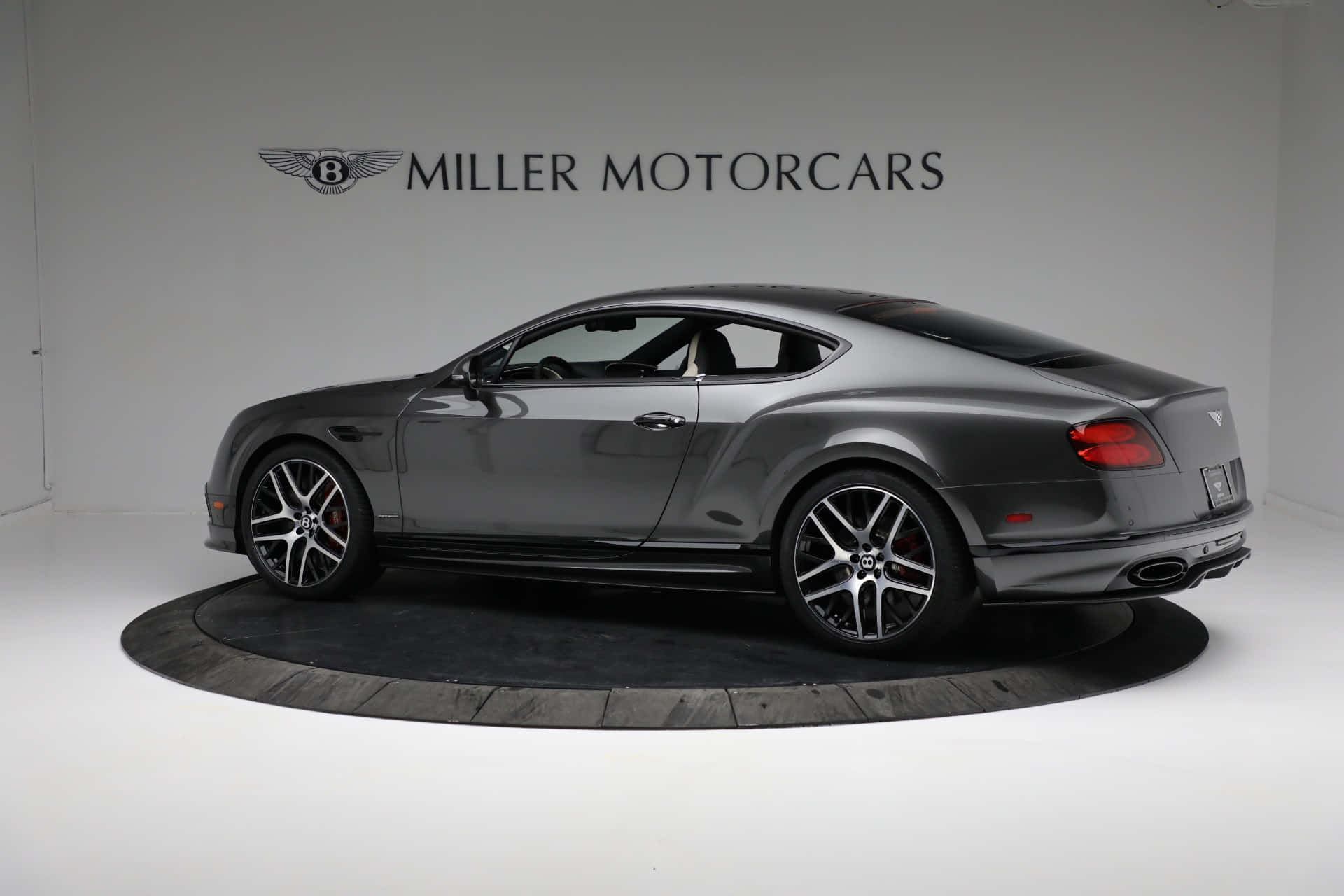 Bentley Continental Supersports 桌布 壁紙 — 一輛時尚的灰色賓利 Continental GT 雙門跑車，在極簡白色工作室的圓形平台上展示，帶有 Miller Motorcars 品牌標識。
