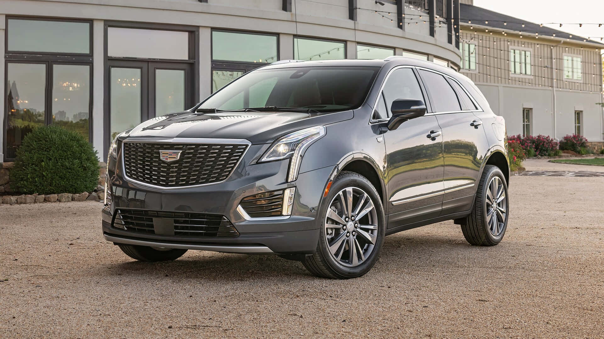 Cadillac Xt5 桌布 壁紙 — 一輛時尚的灰色凱迪拉克SUV停在碎石路上，前方是帶有大窗戶和燈串的現代建築。