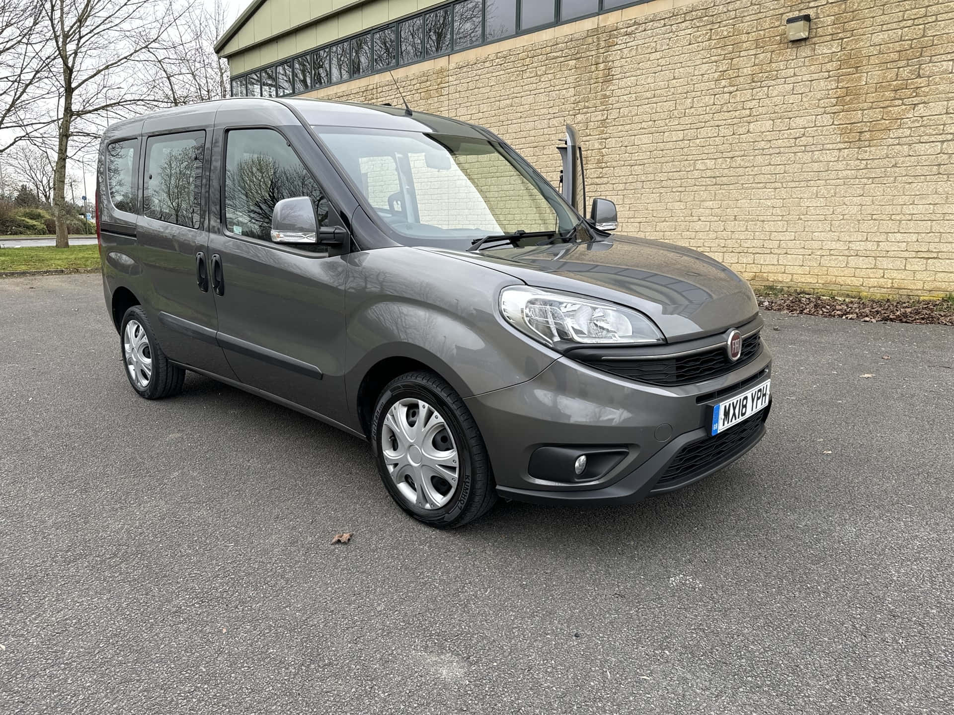  壁紙 — 一架時尚的灰色 Fiat Fiorino 廂型車停放在柏油路上，旁邊是米色磚造建築，背景有光禿禿的樹木。