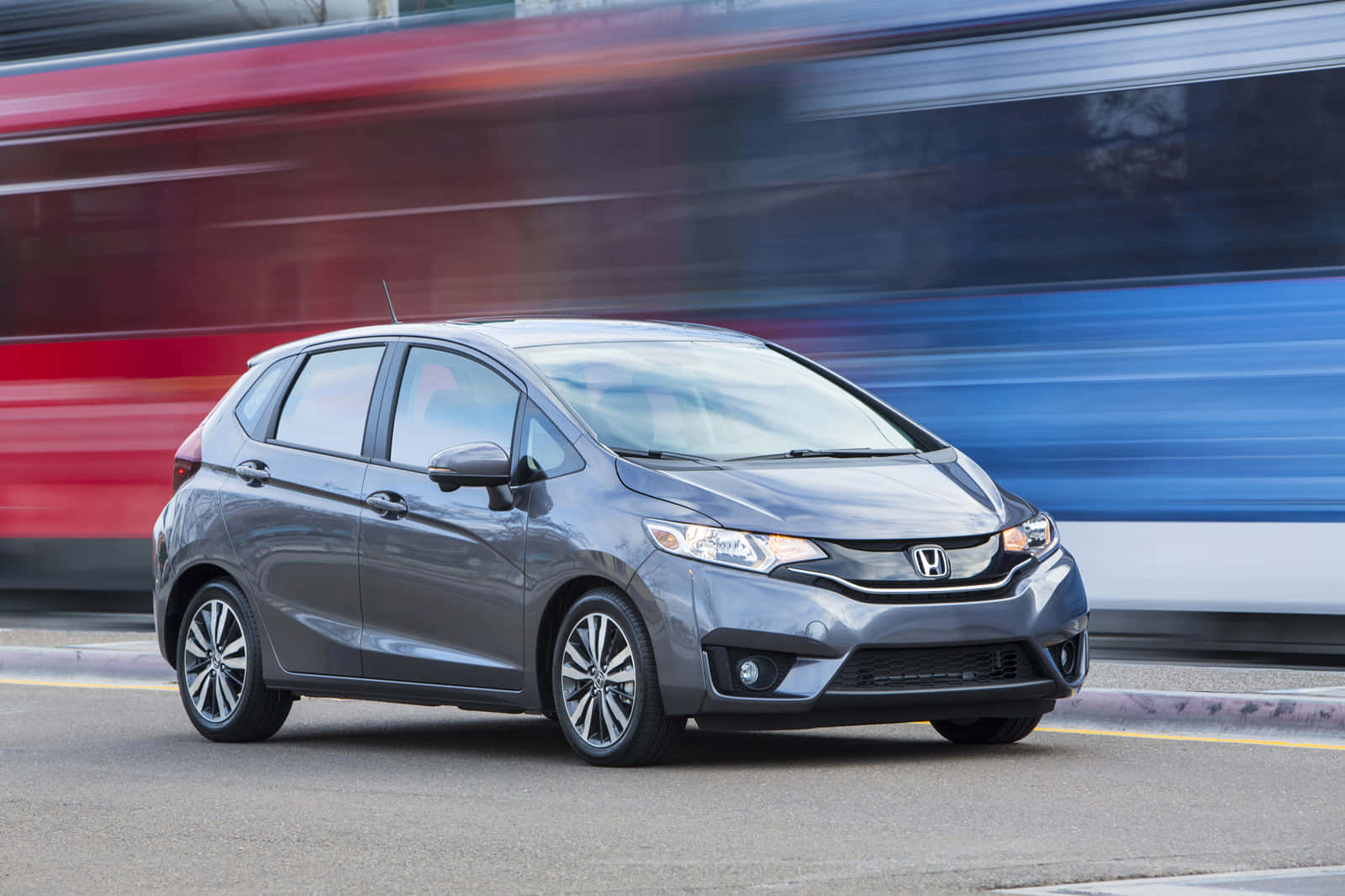 Honda Fit 桌布 壁紙 — 一輛時尚的灰色本田飛度在城市街道上疾馳，身後留下模糊的紅色和藍色運動條紋。