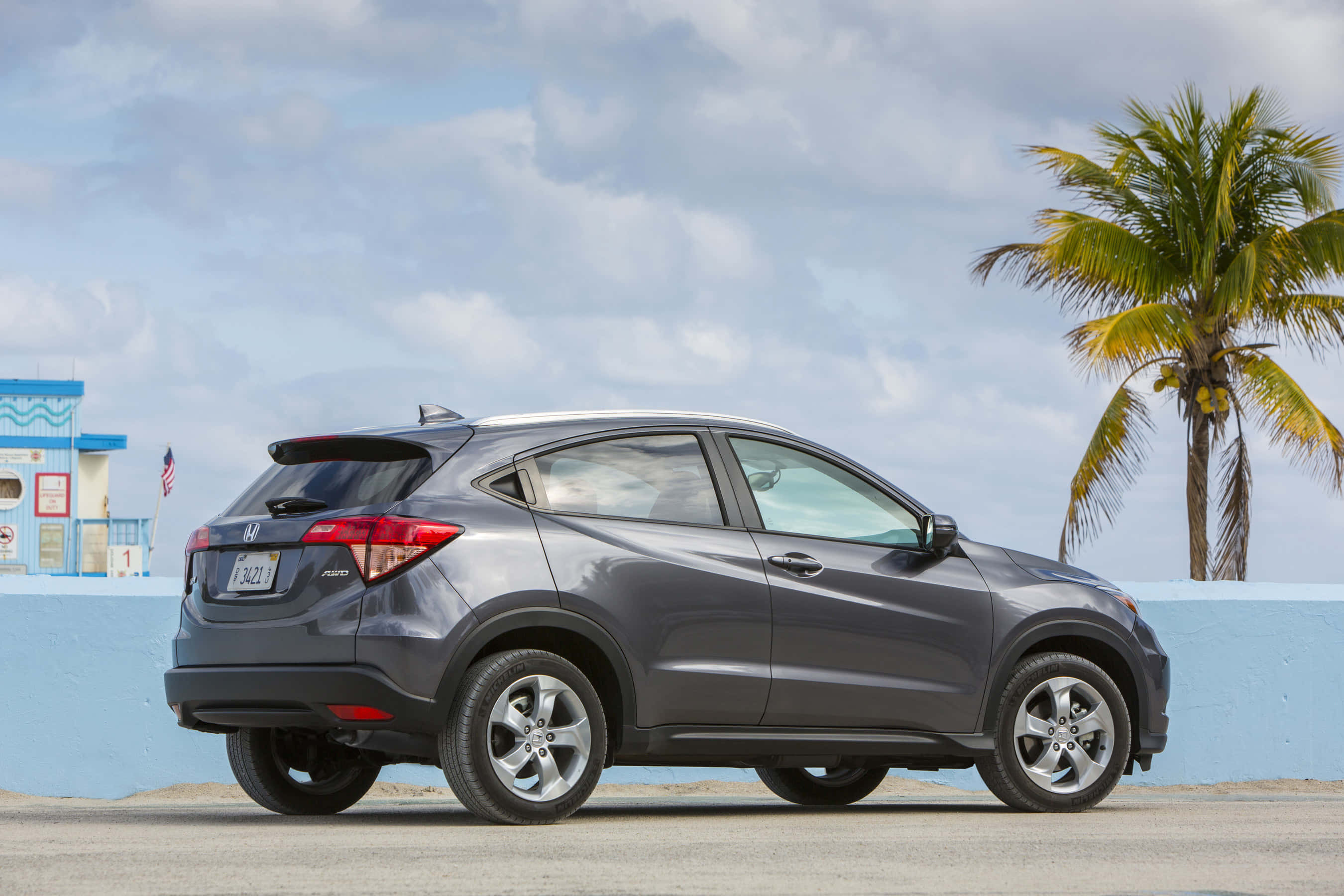 Honda Hr-v 桌布 壁紙 — 一輛時尚的灰色本田 HR-V 停放在棕櫚樹旁，天空局部多雲，靠近一棟藍色建築物。