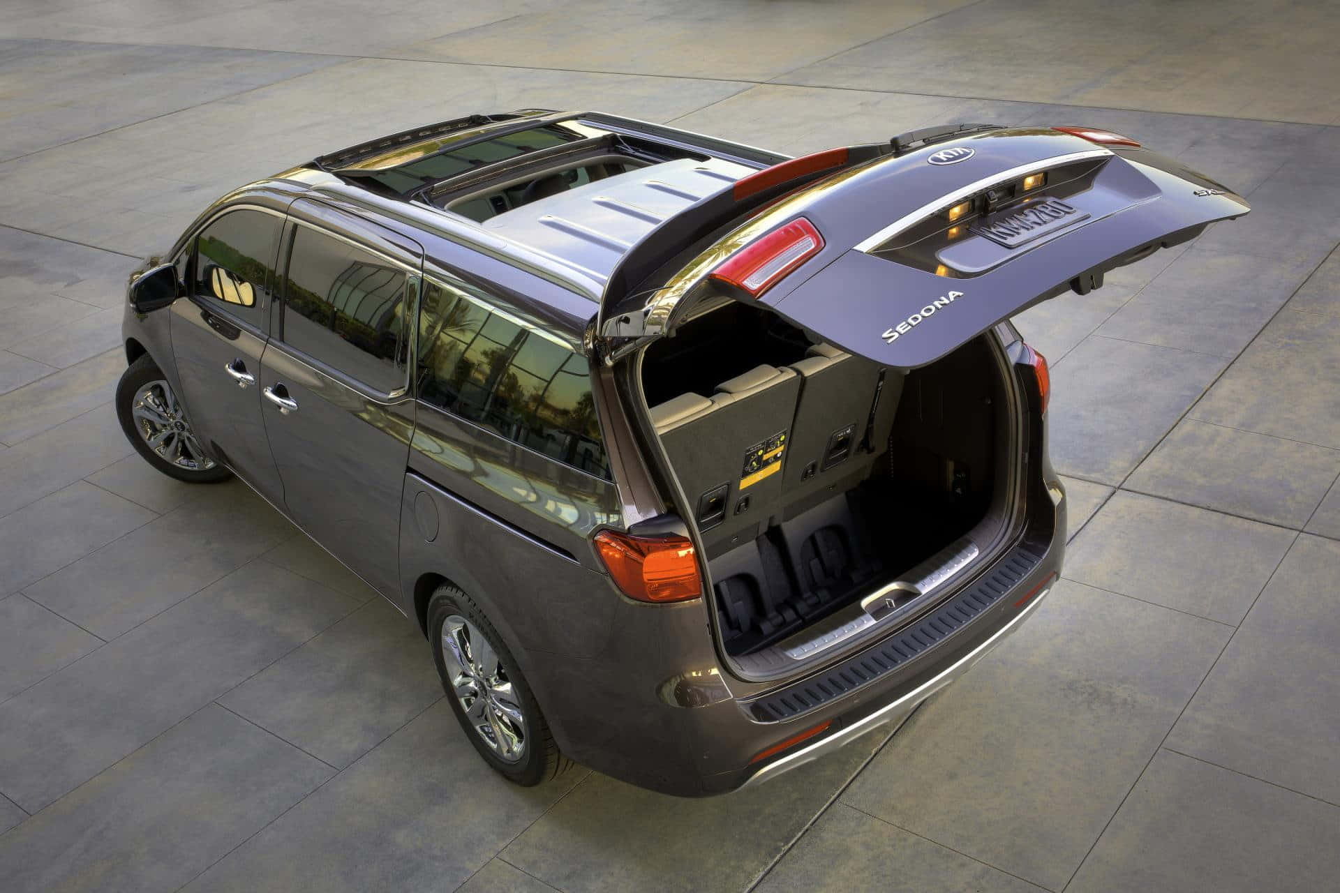 Kia Sedona 桌布 壁紙 — 一輛時尚的灰色Kia Sedona小型貨車，尾門打開，露出鋪磚路面上的寬敞貨物空間。