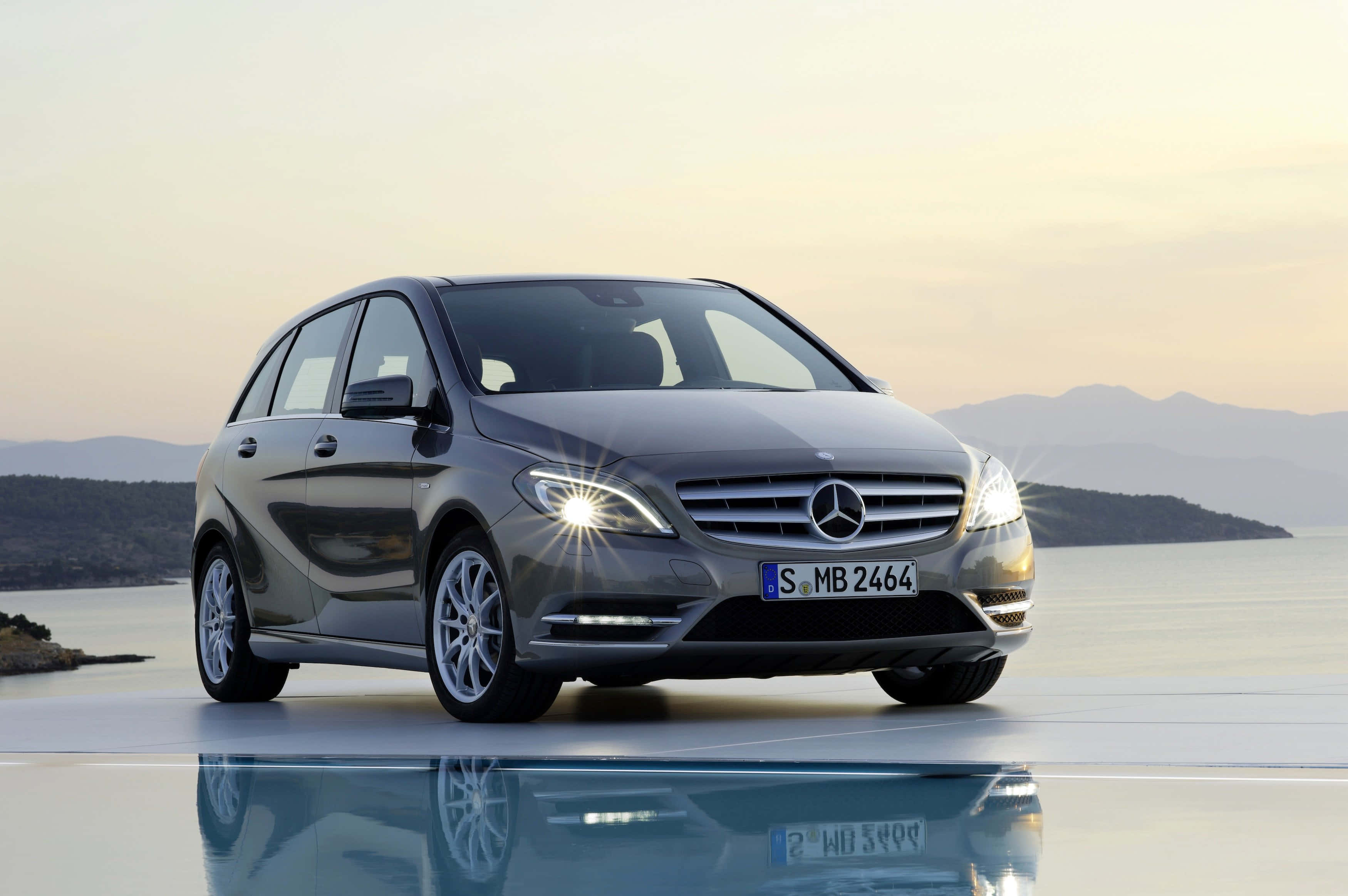 Mercedes Benz B-class 桌布 壁紙 — 一輛流線型灰色 Mercedes-Benz 旅行車在夕陽柔和的金色光芒中，於反射表面上閃耀，背景有山有水。