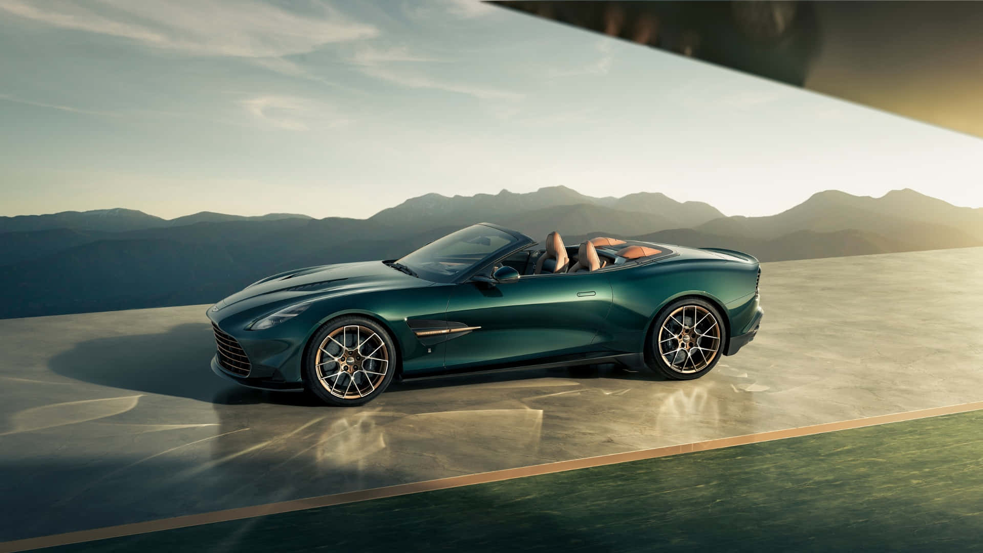  壁紙 — 一輛時尚的綠色Aston Martin DB12 Volante敞篷跑車停放在陽光普照的混凝土平台上，背景是山脈。