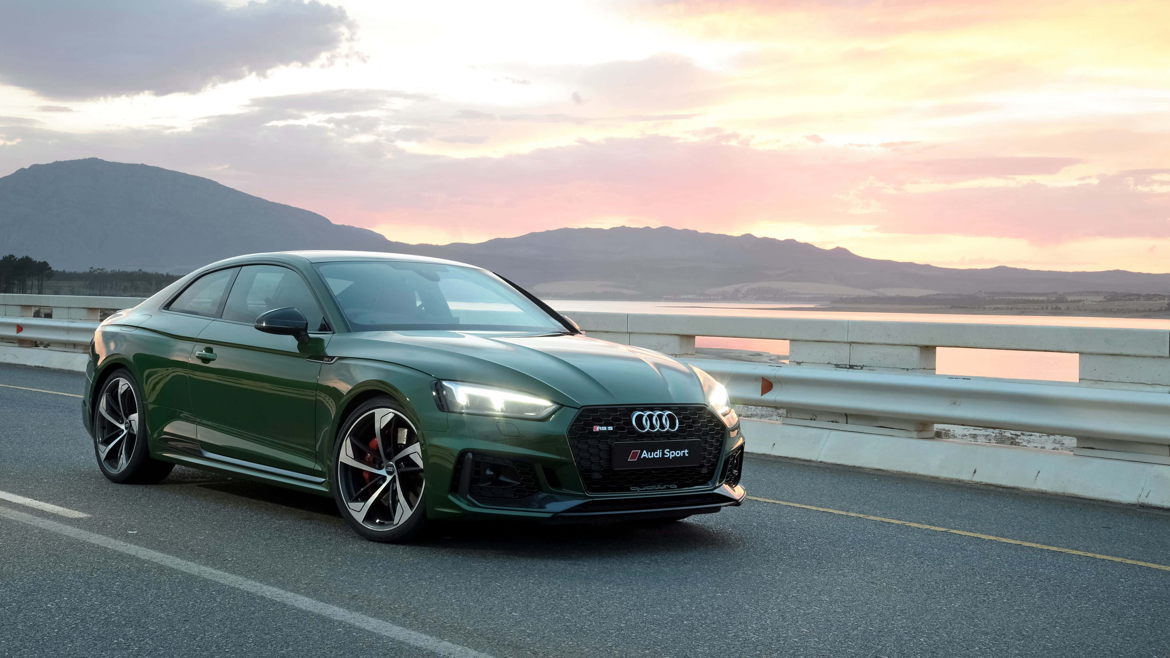 Audi S5 桌布 壁紙 — 一輛流線型綠色奧迪運動轎跑車在日落時分沿著海岸公路行駛，背景是山脈。