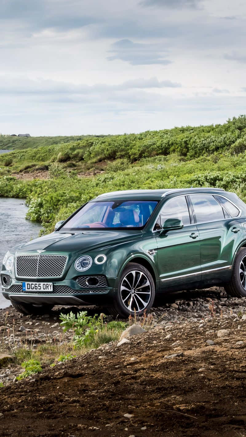 Bentley Bentayga 桌布 壁紙 — 一輛時尚的綠色賓利Bentayga SUV停泊在岩石河邊，背景是茂密的綠色山丘，天空多雲。