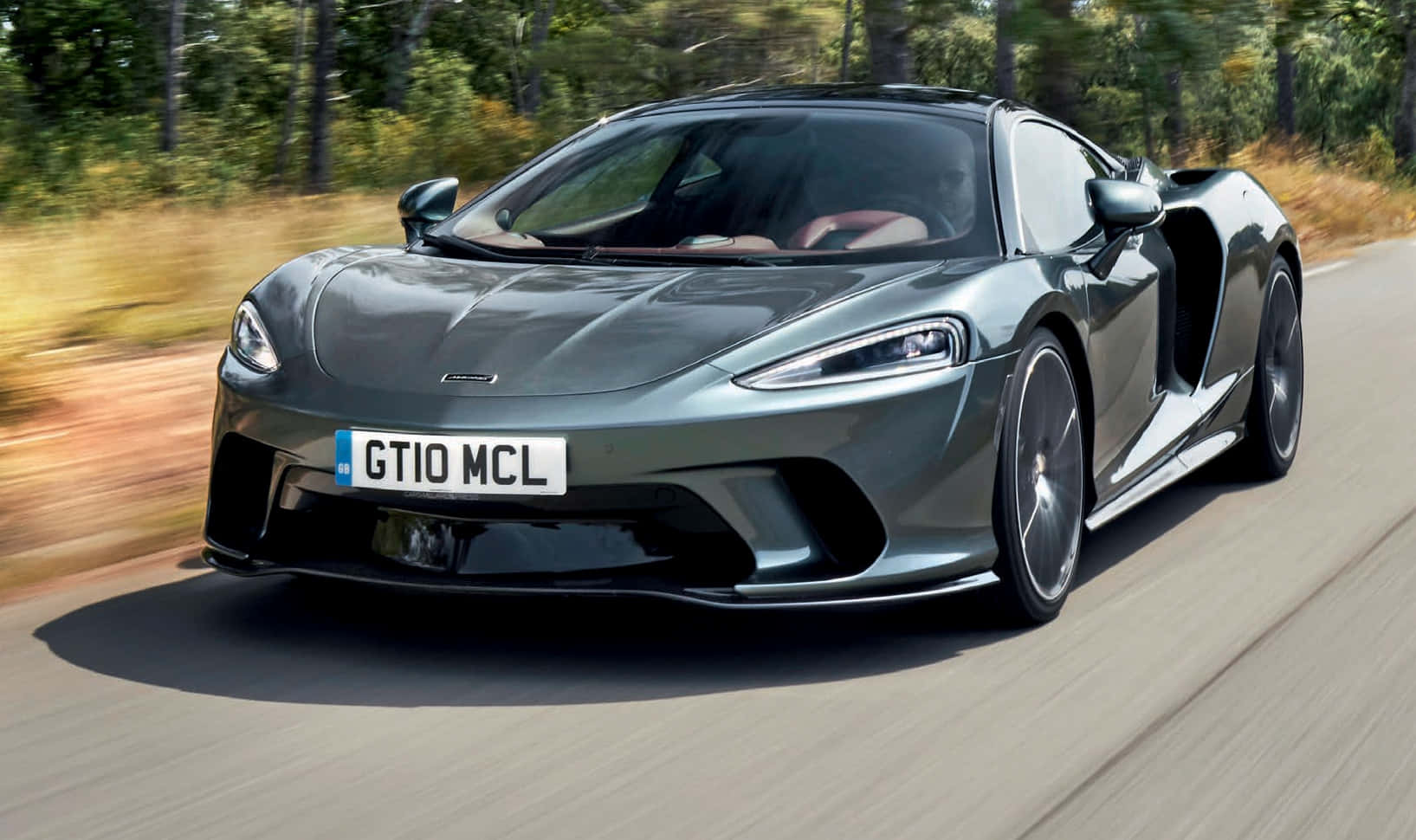 Mclaren Gt 桌布 壁紙 — 一輛時尚的灰色McLaren跑車在森林道路上行駛，動態模糊強調其速度。