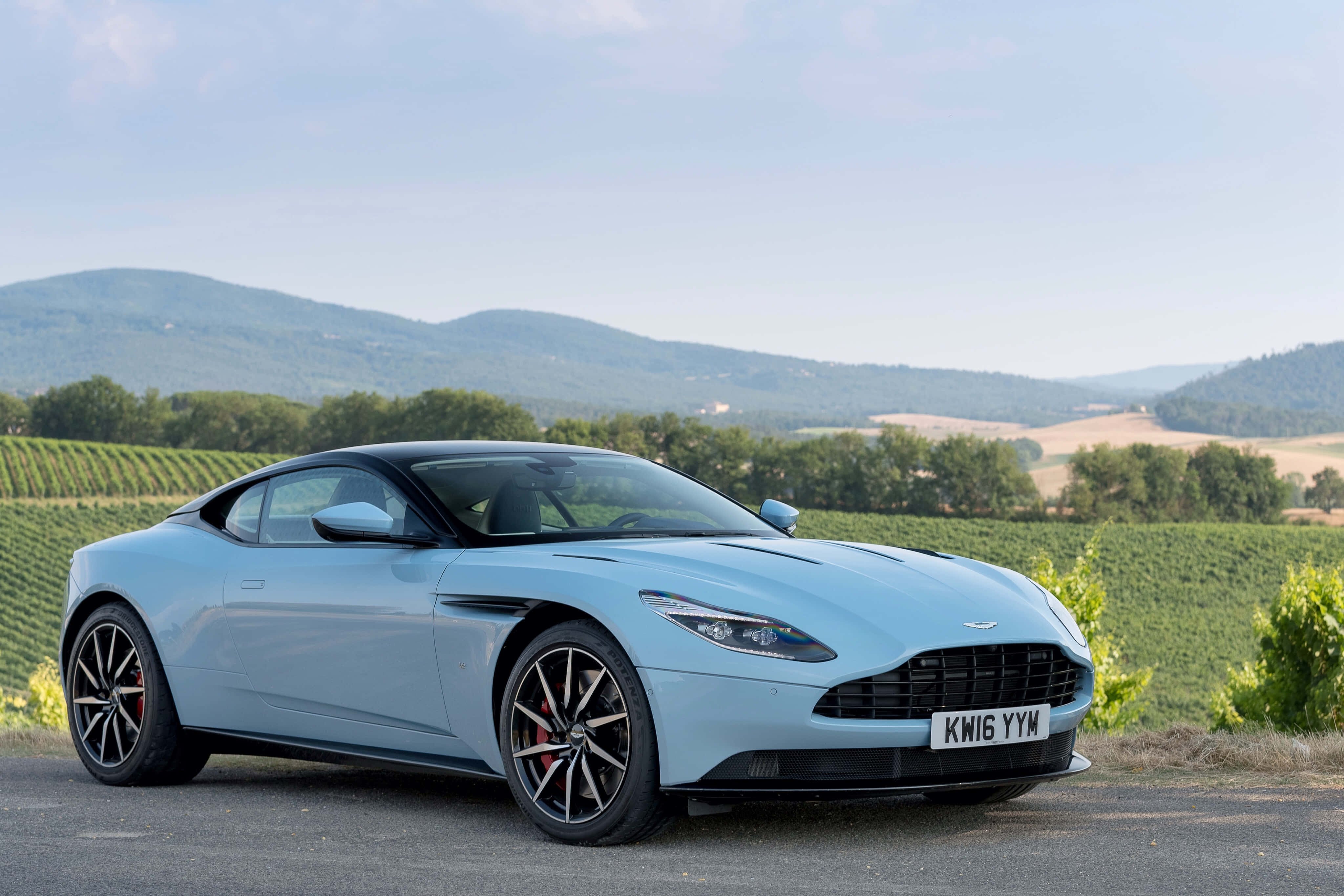 Aston Martin Db11 桌布 壁紙 — 一輛時尚的淺藍色Aston Martin跑車停泊在道路上，背景是起伏的葡萄園山丘和遠處的山脈，天空晴朗。