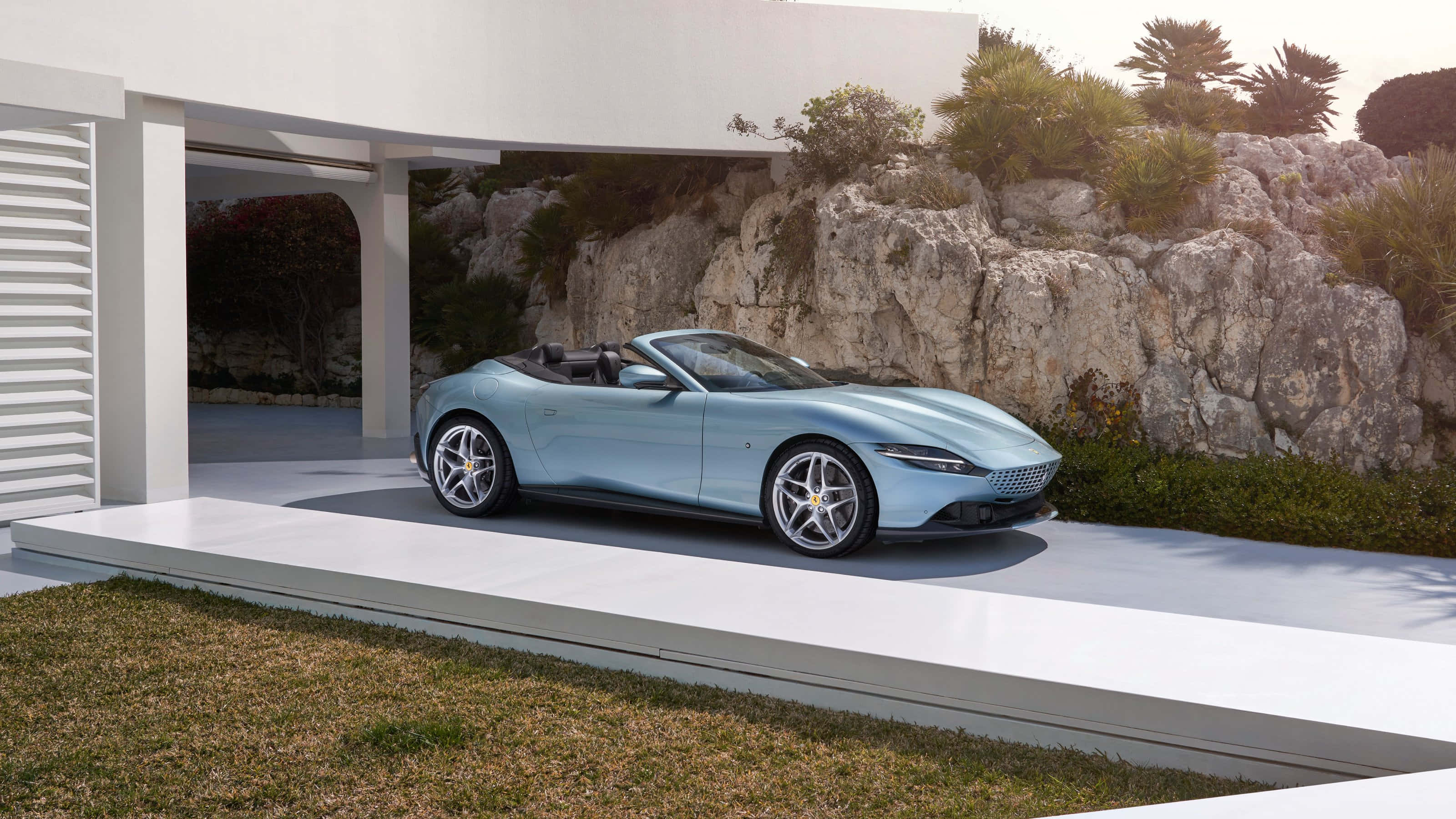 Ferrari Roma 桌布 壁紙 — 一輛時尚淺藍色敞篷跑車停放在現代白色平台上，旁邊是簡約風格的房屋，背景有岩石地形和綠植。