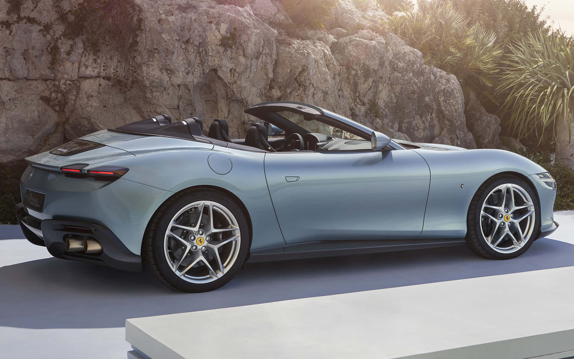 Ferrari Roma 桌布 壁紙 — 一架時尚的淺藍色法拉利敞篷車停放在現代化平台上，背景是陽光普照的岩石懸崖和棕櫚樹。