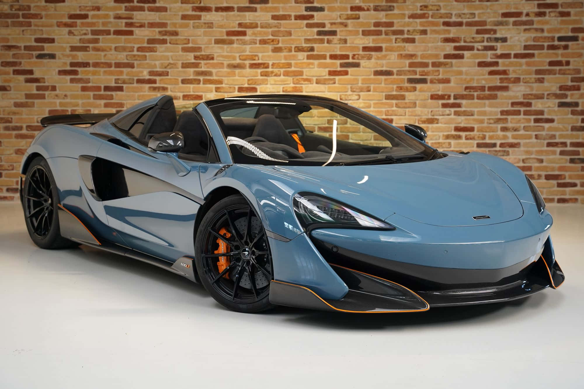 Mclaren 600lt Spider 桌布 壁紙 — 一架時尚的淺藍色McLaren跑車帶有橙色點綴，停放在白色地板上的質樸磚牆旁。