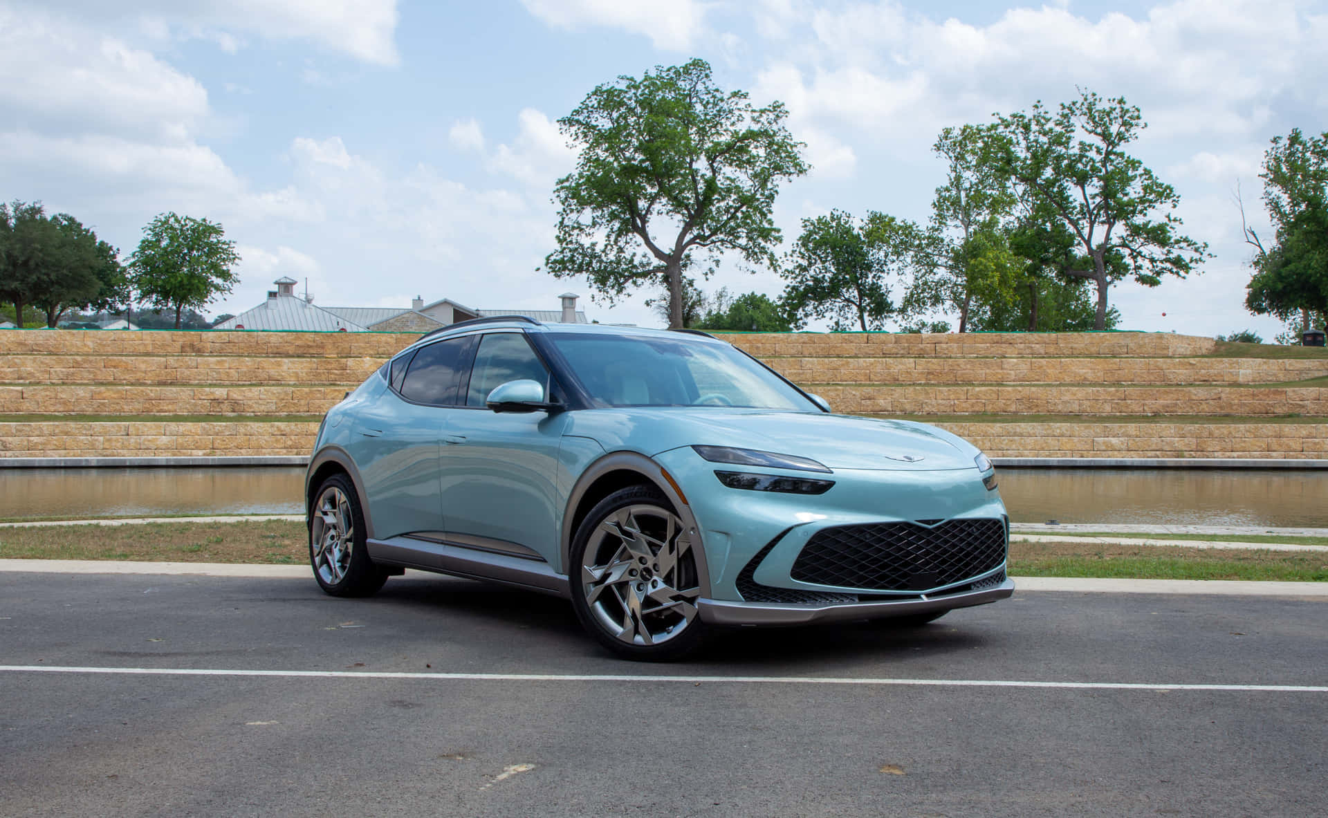 Genesis Gv60 桌布 壁紙 — 一部時尚的淺藍色現代SUV停放在柏油路上，旁邊有石牆和水景，天空部分多雲。