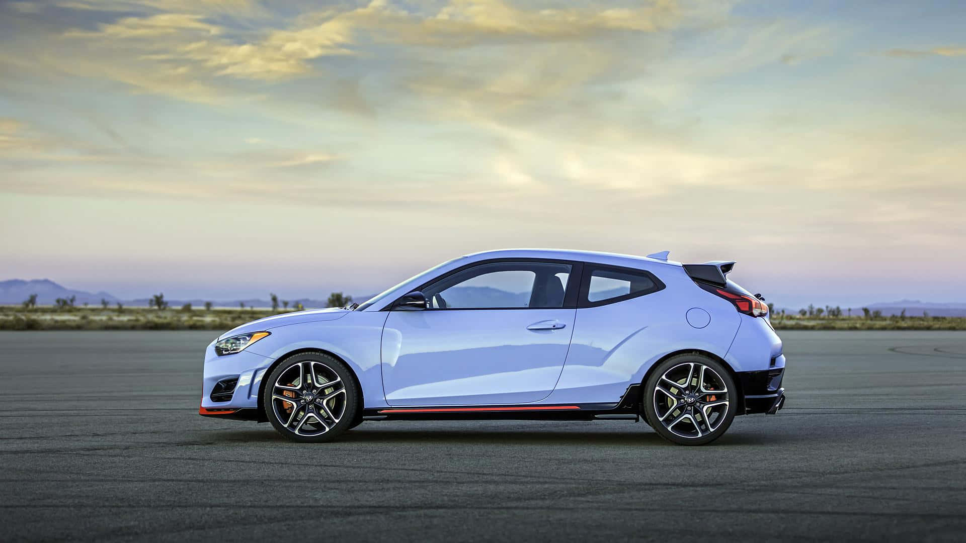 Hyundai Veloster 桌布 壁紙 — 一輛時尚的淺藍色跑車，帶有黑色飾邊和紅色裝飾，在柔和的金色日落天空下，停放在廣闊的瀝青賽道上。