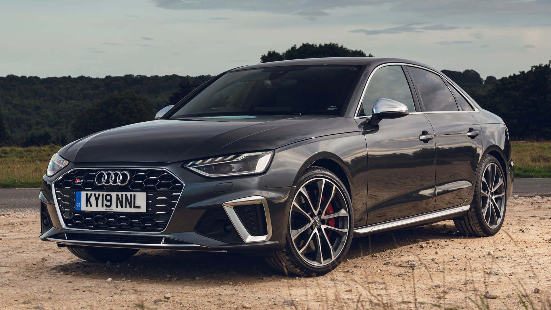 Audi S4 桌布 壁紙 — 一輛時尚的啞光黑色奧迪RS4轎車停泊在碎石路上，周圍是森林覆蓋的山丘，天空多雲。