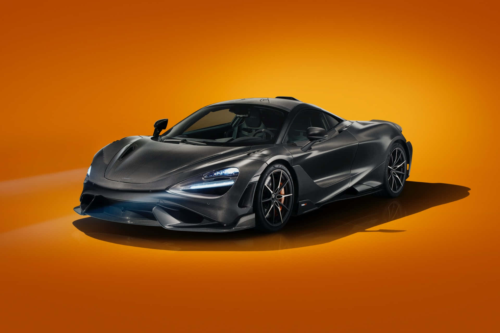 Mclaren 765lt 桌布 壁紙 — 一輛時尚的啞光黑色McLaren跑車在溫暖的橙色工作室燈光下閃耀，其空氣動力學車身上反射出銳利的光澤。