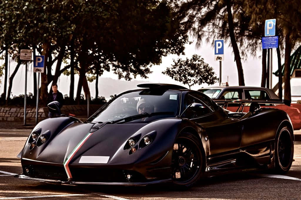 Pagani Zonda Tricolore 桌布 壁紙 — 一架時尚的啞光黑色帕加尼Huayra跑車停泊在林蔭大道上，附近有一個人。