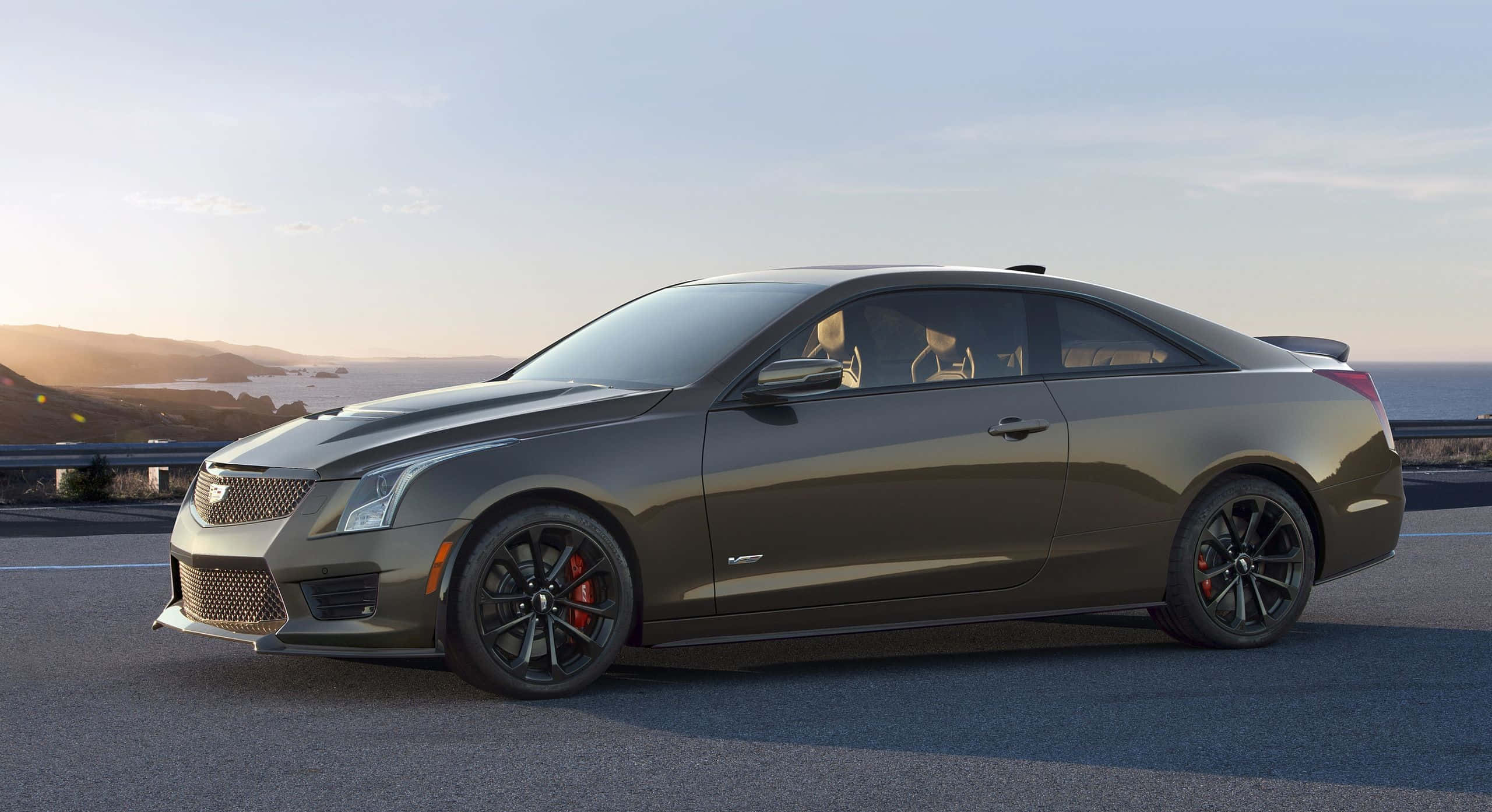 Cadillac Ats 桌布 壁紙 — 一輛時尚的消光灰色Cadillac ATS-V雙門跑車停放在日落時分的海岸公路上，背景是海洋和山丘。