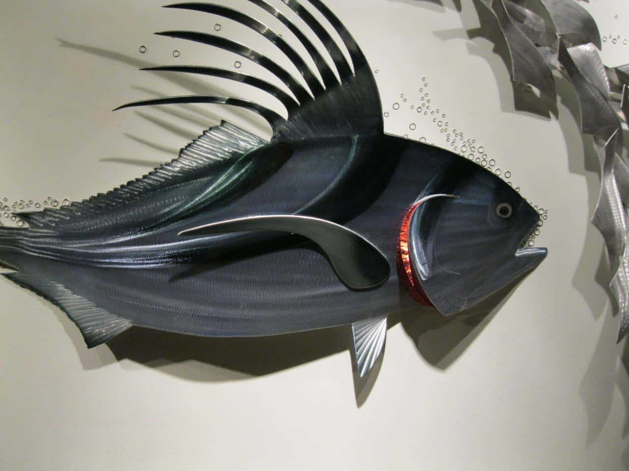 Roosterfish 桌布 壁紙 — 一個光滑的金屬魚雕塑，帶有紅色點綴和氣泡細節，安裝在牆上。
