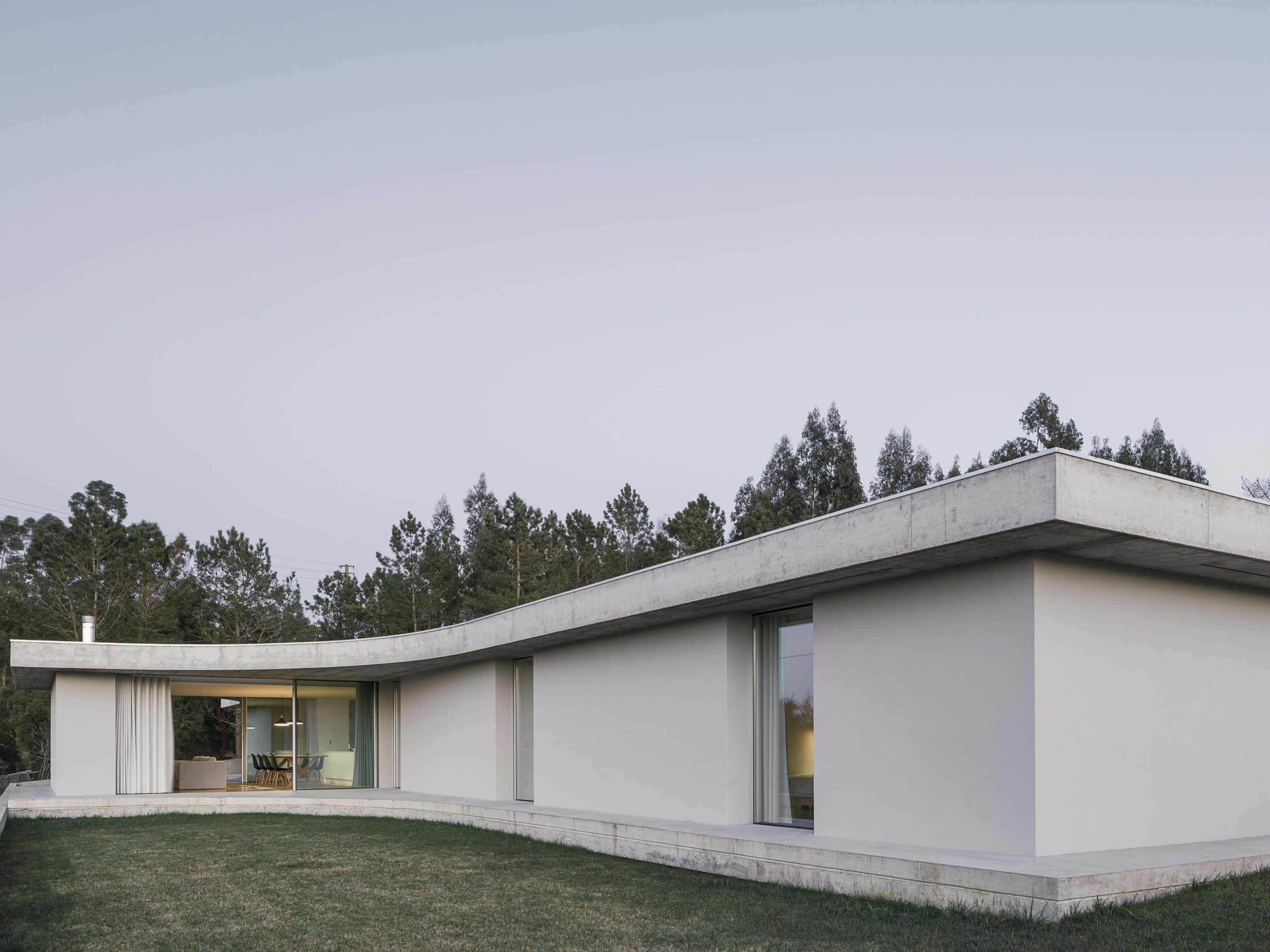 Minimalist Architecture 桌布 壁紙 — 一座時尚的現代混凝土房屋，帶有弧形牆壁和大玻璃門，坐落在草坪上，周圍環繞著高大的松樹，天空蒼白。