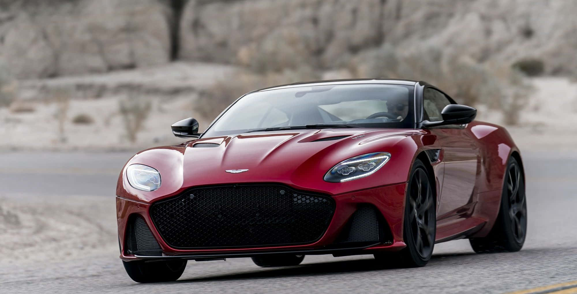 Aston Martin Dbs Superleggera 桌布 壁紙 — 一輛流線型紅色Aston Martin跑車在沙漠公路上行駛，背景是岩石峭壁。