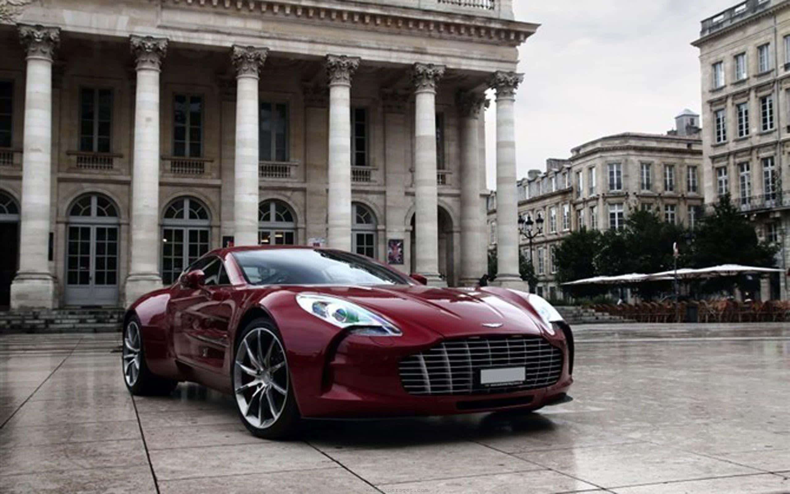 Aston Martin One-77 桌布 壁紙 — 一輛流線型紅色Aston Martin跑車在濕潤的廣場上閃閃發光，前方是擁有高柱的宏偉新古典主義建築。