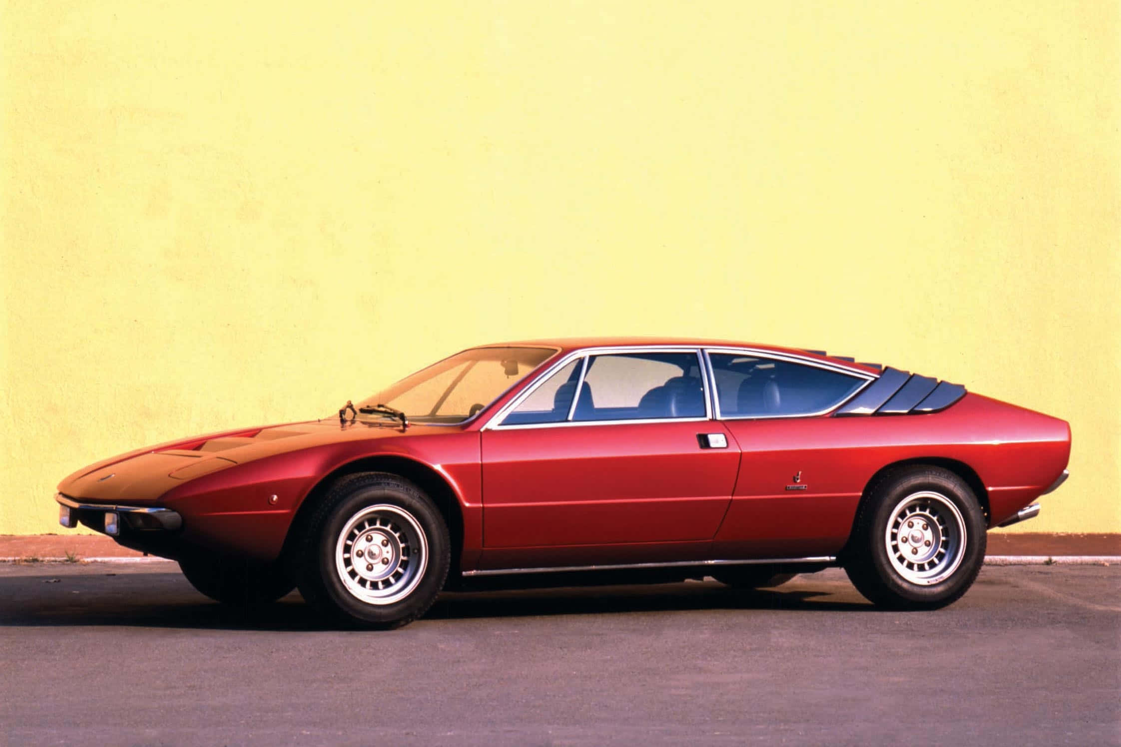 Lamborghini Urraco 桌布 壁紙 — 一輛線條流暢的紅色經典跑車，具有獨特的楔形車身，停靠在純黃色牆壁旁。