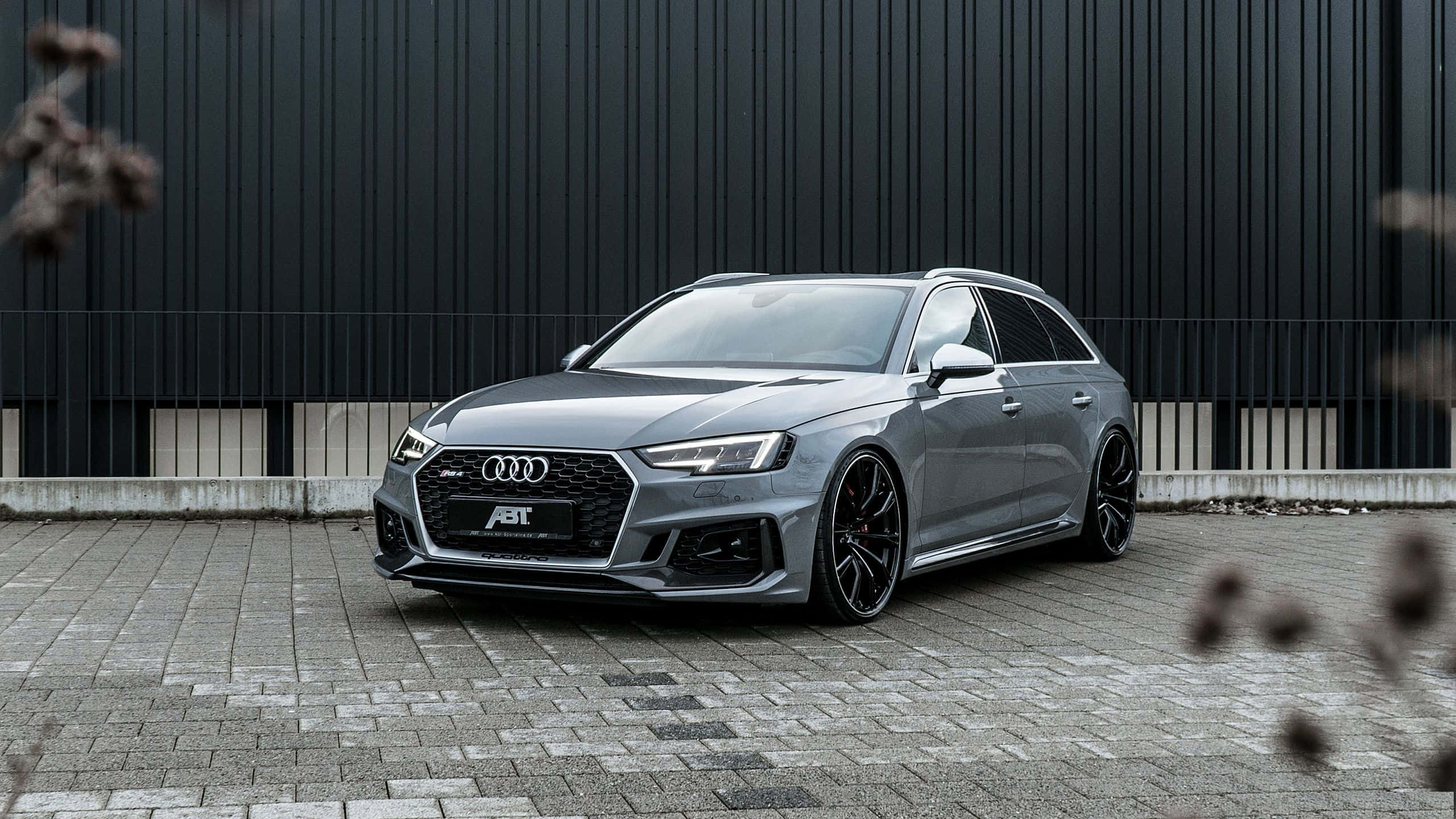 Audi Rs4 桌布 壁紙 — 一架時尚銀色ABT改裝奧迪RS4 Avant旅行車停放在鋪砌的庭院中，背景是深色工業牆。