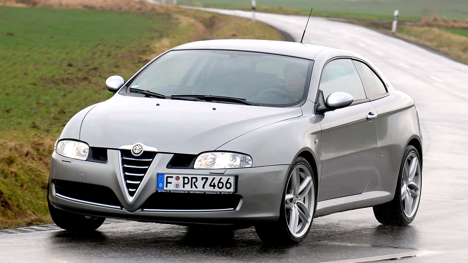 Alfa Romeo Gt 桌布 壁紙 — 一輛時尚的銀色愛快羅密歐雙門跑車在陰天下行駛在濕潤的鄉村道路上，兩旁是綠色的田野。