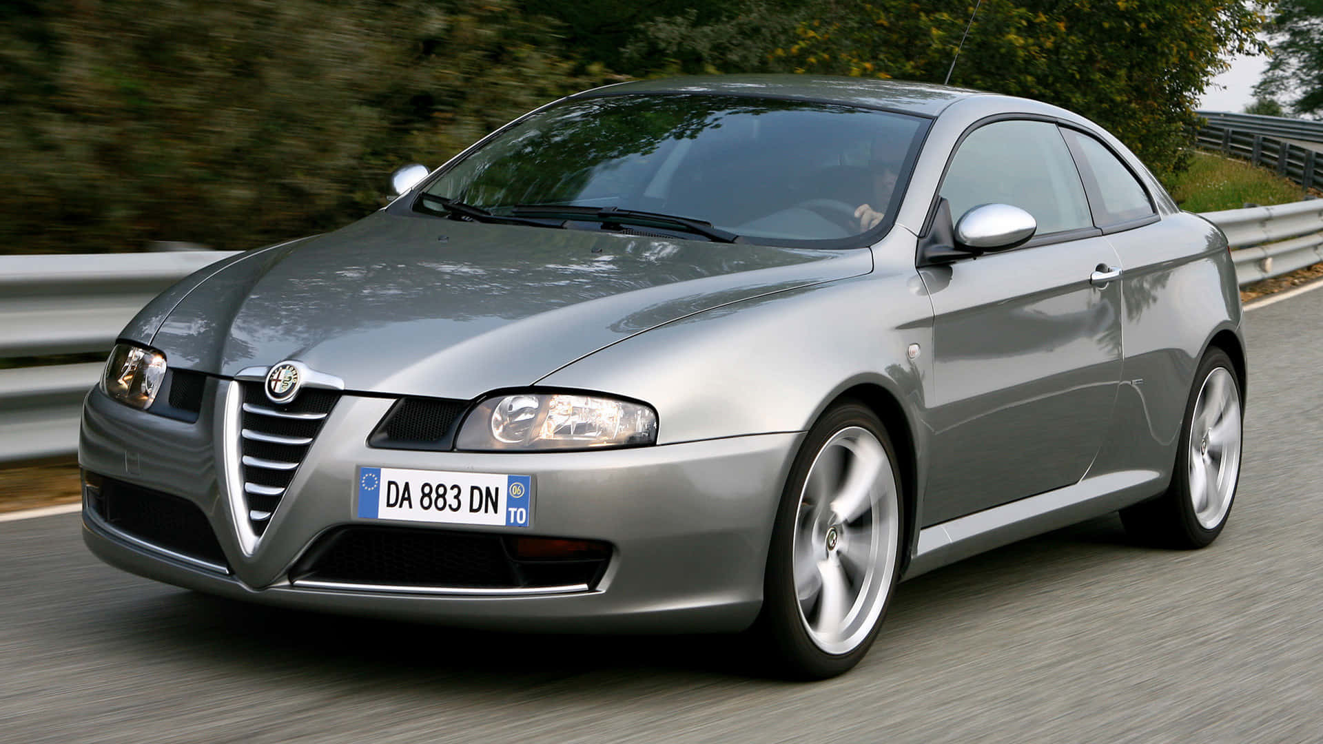 Alfa Romeo Gt 桌布 壁紙 — 一輛時尚的銀色愛快羅密歐雙門跑車沿著蜿蜒的道路疾馳，綠意和護欄模糊移動。