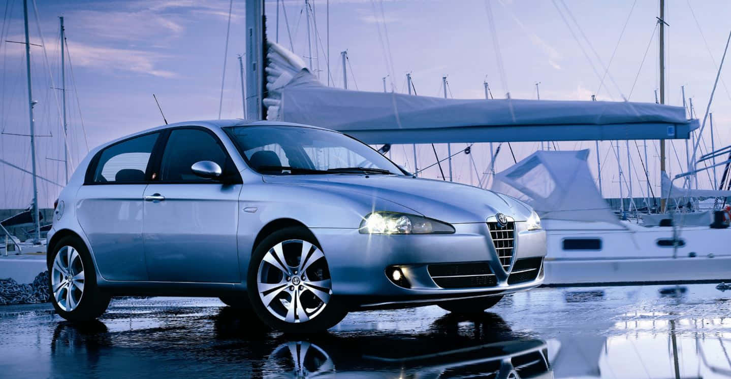 Alfa Romeo 147 桌布 壁紙 — 一輛時尚的銀色愛快羅密歐掀背車在暮色天空下的濕滑碼頭旁，在帆船旁邊閃閃發光。