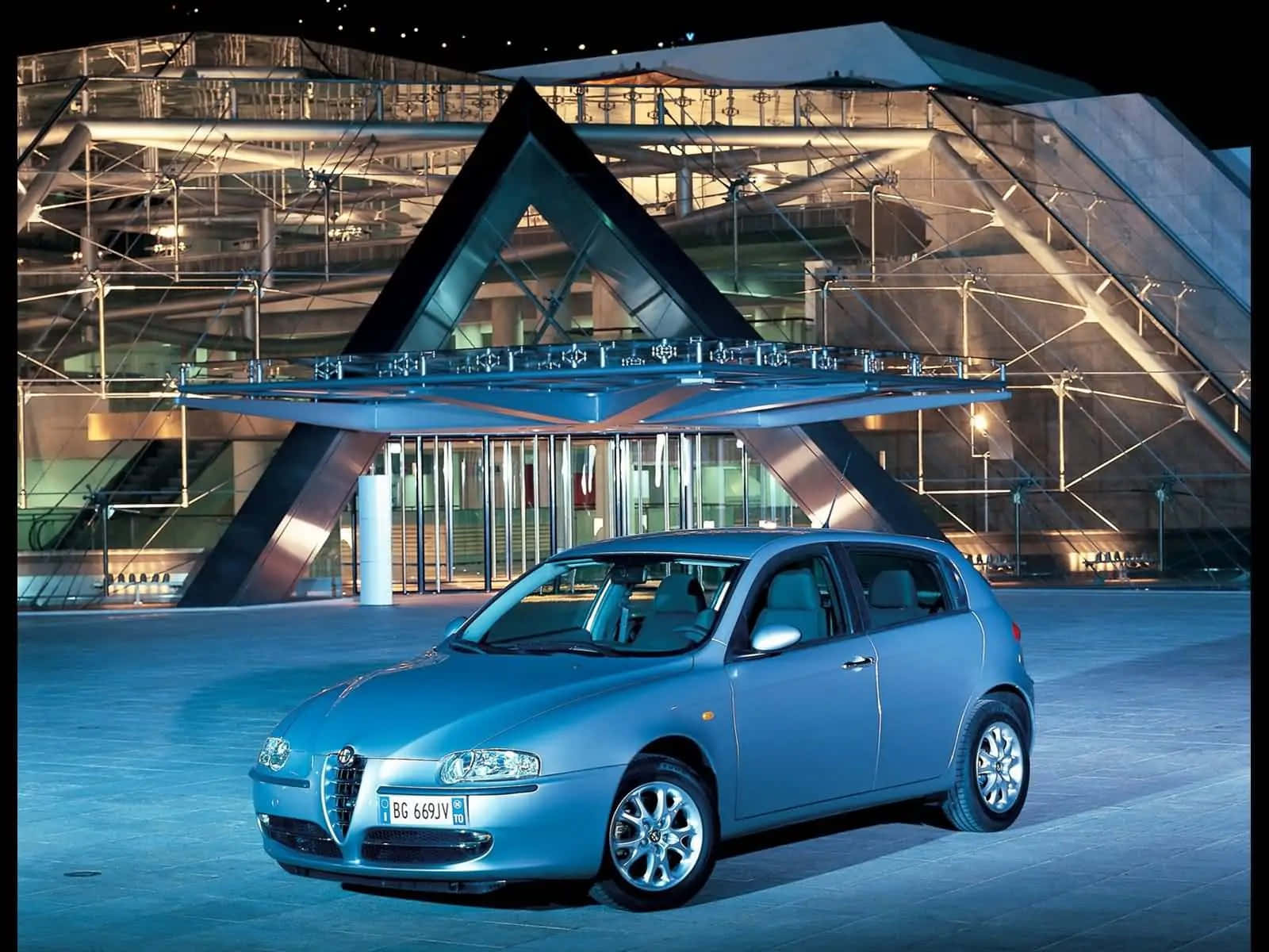 Alfa Romeo 147 桌布 壁紙 — 一輛時尚的銀色Alfa Romeo掀背車在夜晚停泊在現代玻璃和鋼結構建築前。