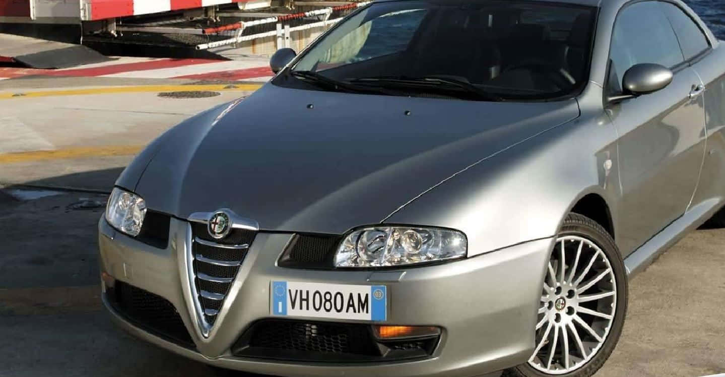 Alfa Romeo Gt 桌布 壁紙 — 一台時尚的銀色愛快羅密歐跑車雙門轎跑車停放在混凝土場地上，背景有紅白條紋屏障。