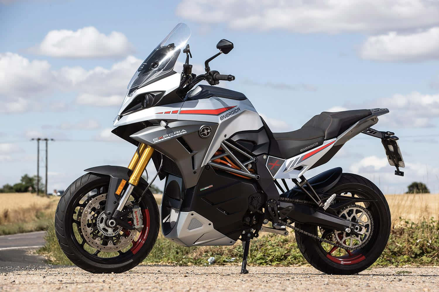  壁紙 — 一架時尚的銀黑色 Energica 摩托車，配金色前叉，停放在碎石路上，天空局部多雲。