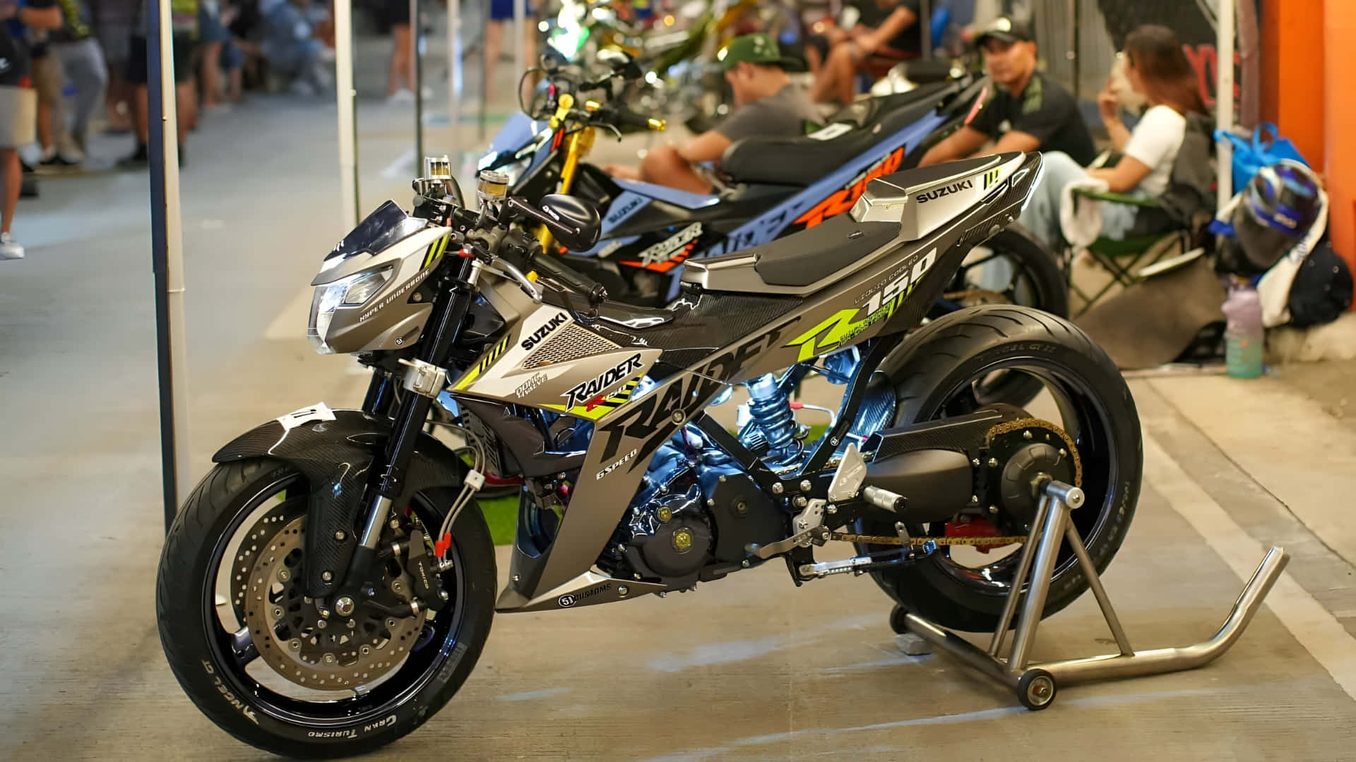  壁紙 — 一輛時尚的銀黑色鈴木 Raider R150 機車，放在繁忙的室內展覽廳的支架上。