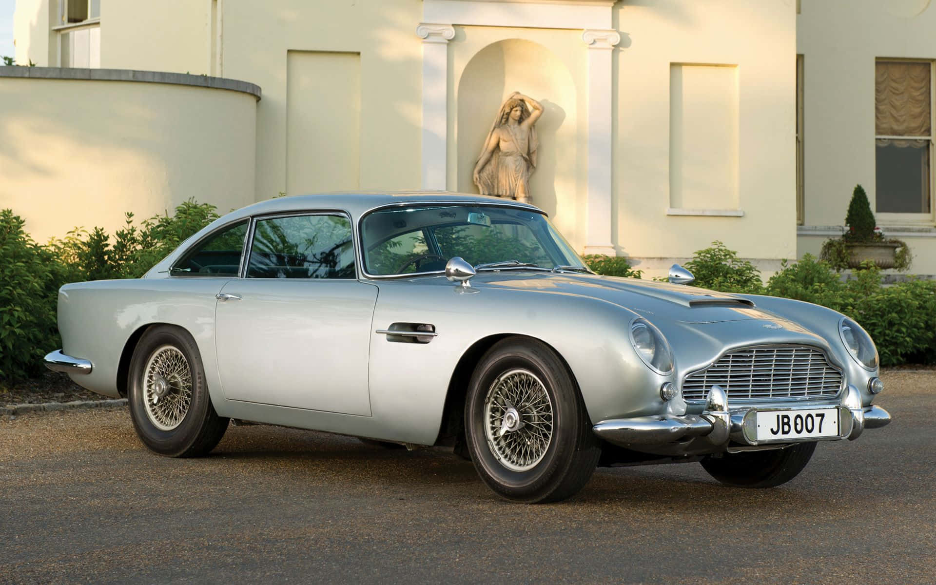 Aston Martin Db5 桌布 壁紙 — 一架流線型銀色Aston Martin DB5優雅地停放在一棟帶有古典雕像的宏偉奶油色豪宅前。