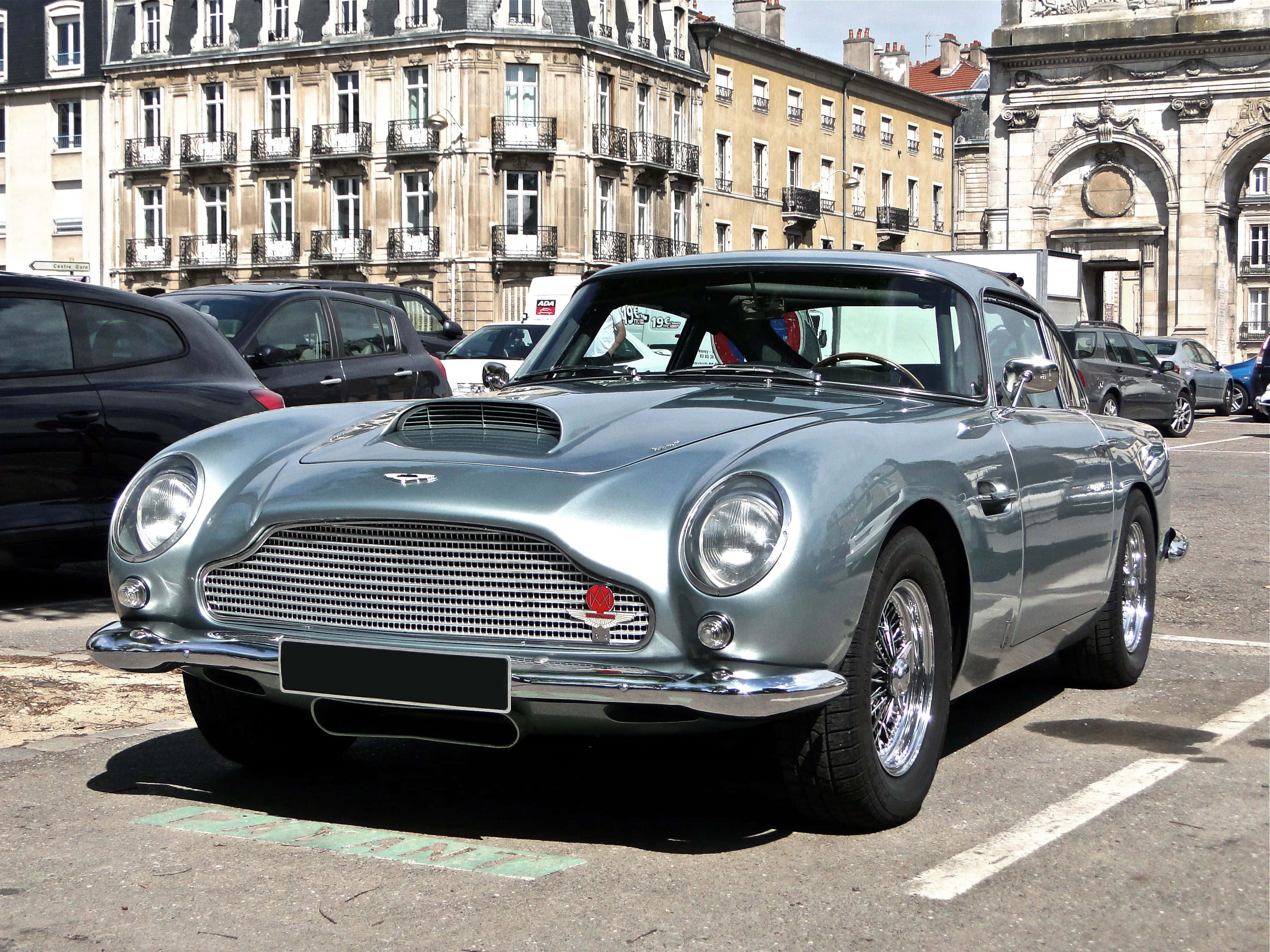 Aston Martin Db5 桌布 壁紙 — 一架時尚的銀色Aston Martin DB6停放在陽光普照的歐洲城市廣場，背後是歷史建築。