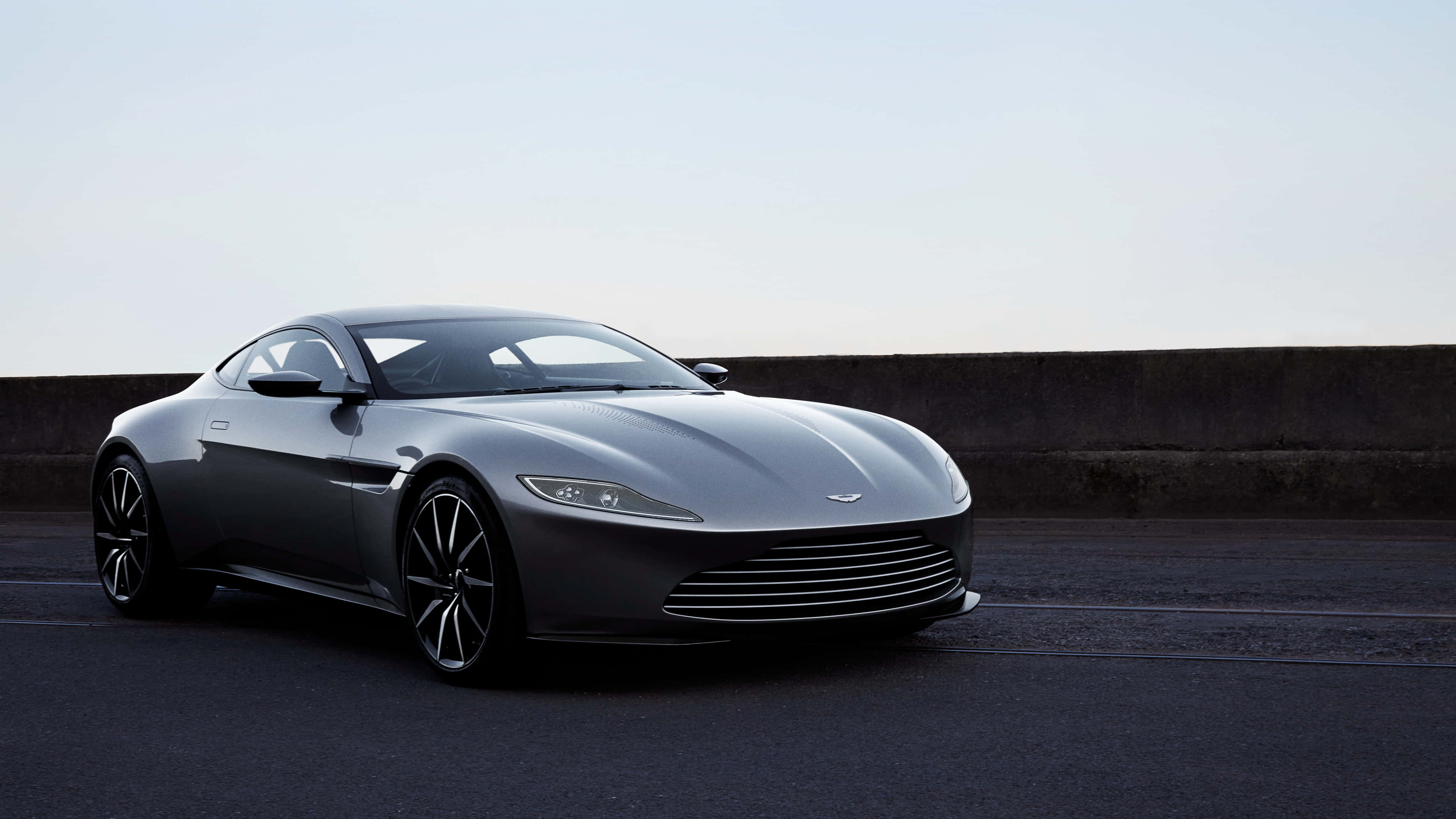 Aston Martin Vantage 桌布 壁紙 — 一架時尚的銀色Aston Martin跑車停泊在昏暗的瀝青路上，旁邊是混凝土護欄，天空蒼白。