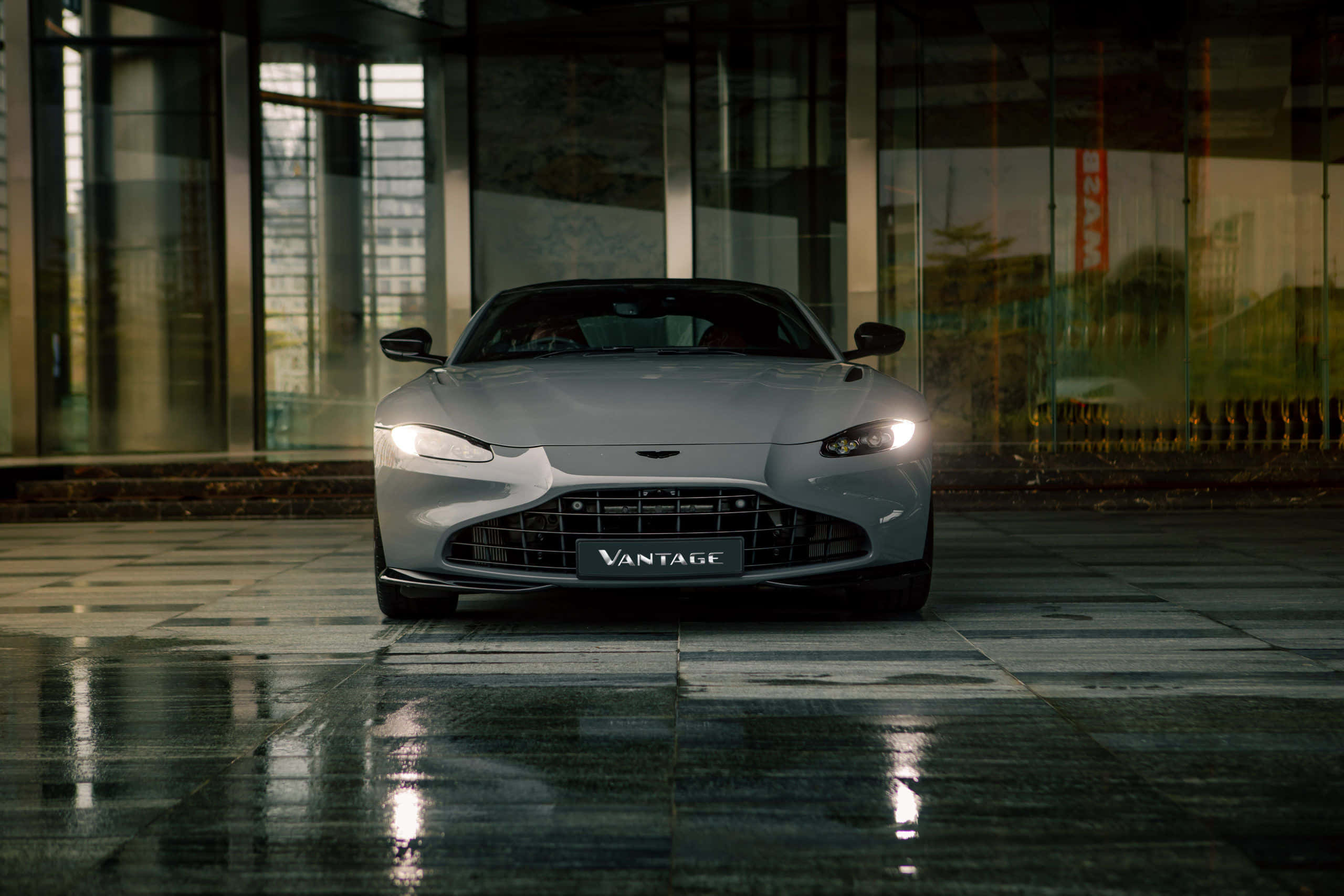 Aston Martin Vantage 桌布 壁紙 — 一輛流線型的銀色Aston Martin Vantage在夜間於玻璃建築旁的濕瓷磚廣場上，在車頭燈下閃閃發光。