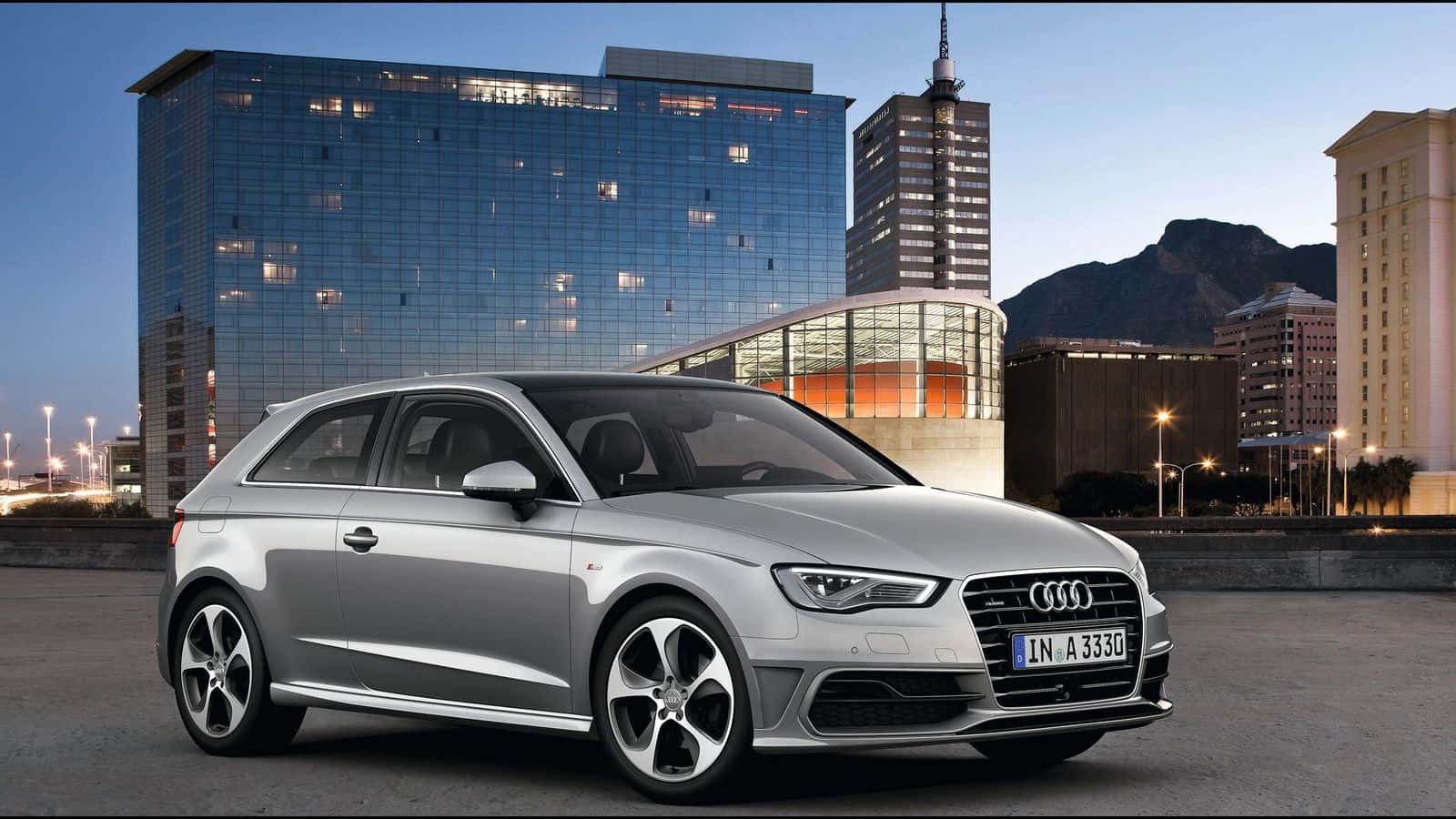 Audi A3 桌布 壁紙 — 一輛時尚的銀色奧迪A3 Sportback停放在現代玻璃摩天大樓和山脈前，正值黃昏。