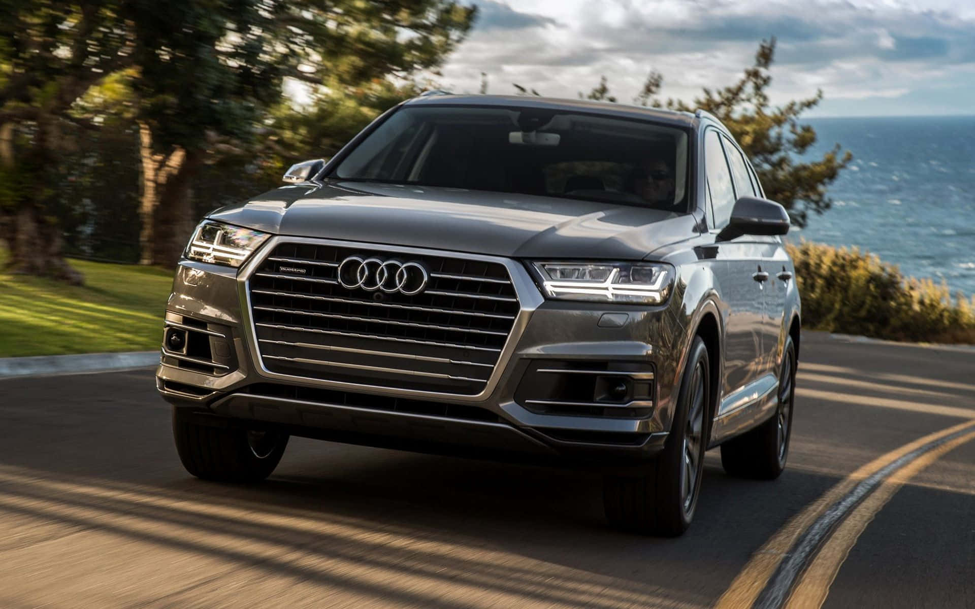 Audi Q7 桌布 壁紙 — 一輛時尚的銀色奧迪Q7 SUV沿著海岸道路行駛，背景是海洋和樹木，天空多雲。