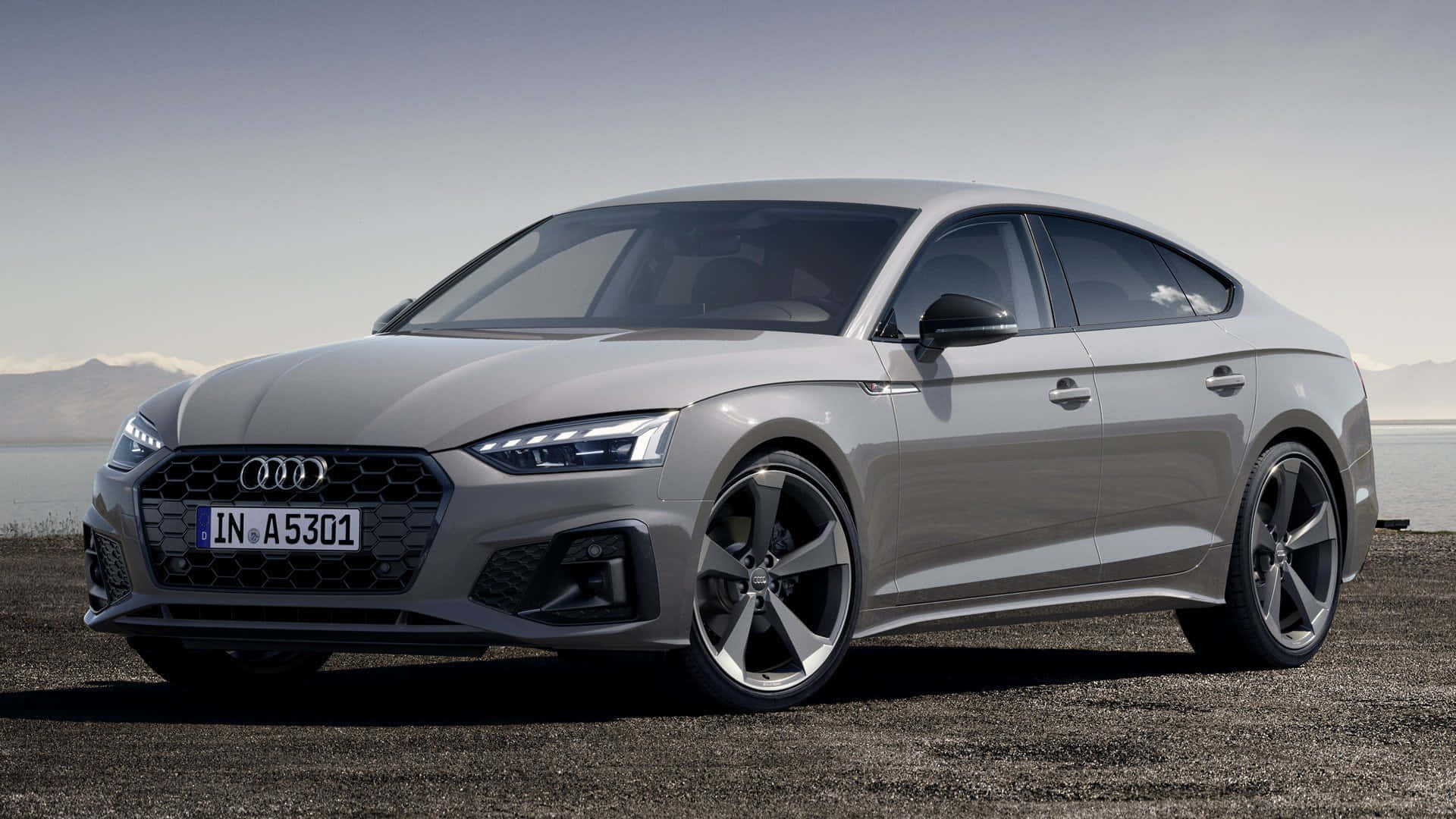 Audi A5 桌布 壁紙 — 一輛時尚的銀色奧迪轎車停泊在碎石上，靠近水邊，遠處有山巒，天空晴朗。