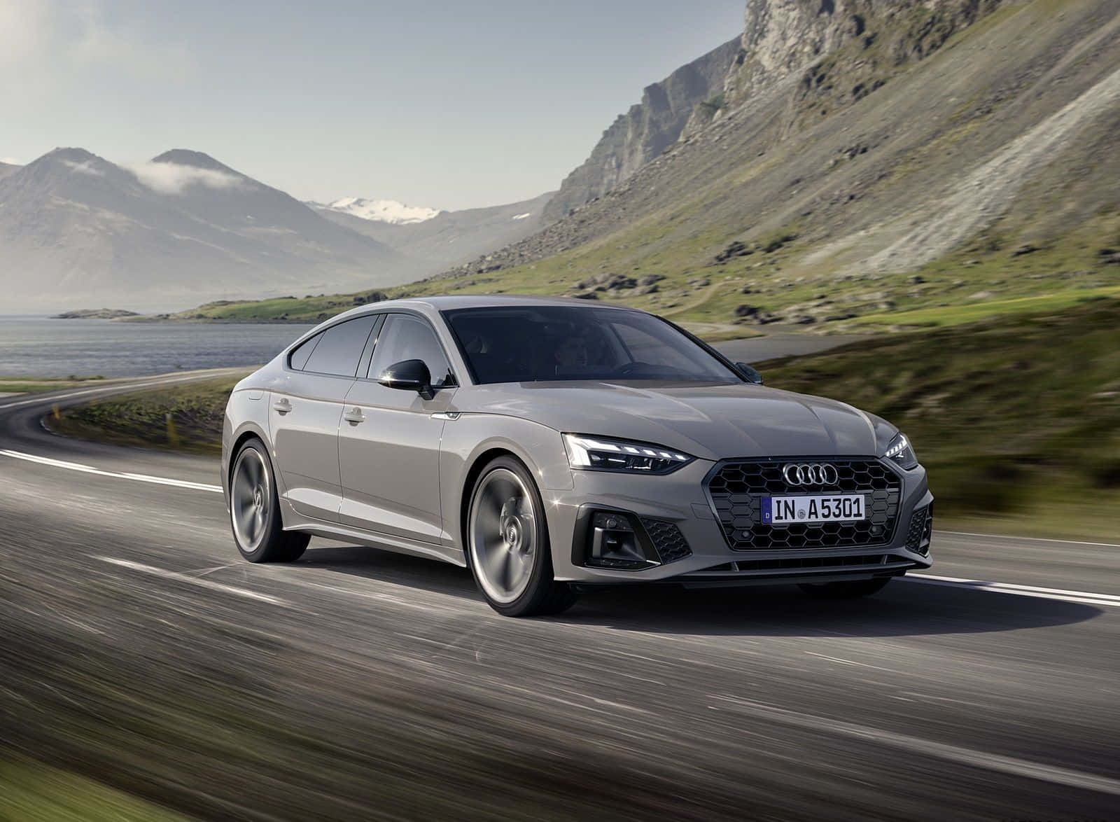Audi A5 桌布 壁紙 — 一輛時尚的銀色奧迪轎車沿著蜿蜒的海岸公路疾馳，兩旁是崎嶇的山脈和寧靜的大海，天空晴朗。