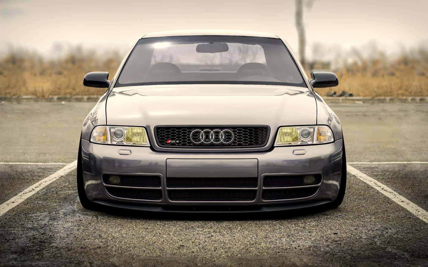 Audi Rs5 桌布 壁紙 — 一輛時尚的銀色奧迪跑車，降低懸吊，停放在空曠的瀝青場地上，天空多雲。