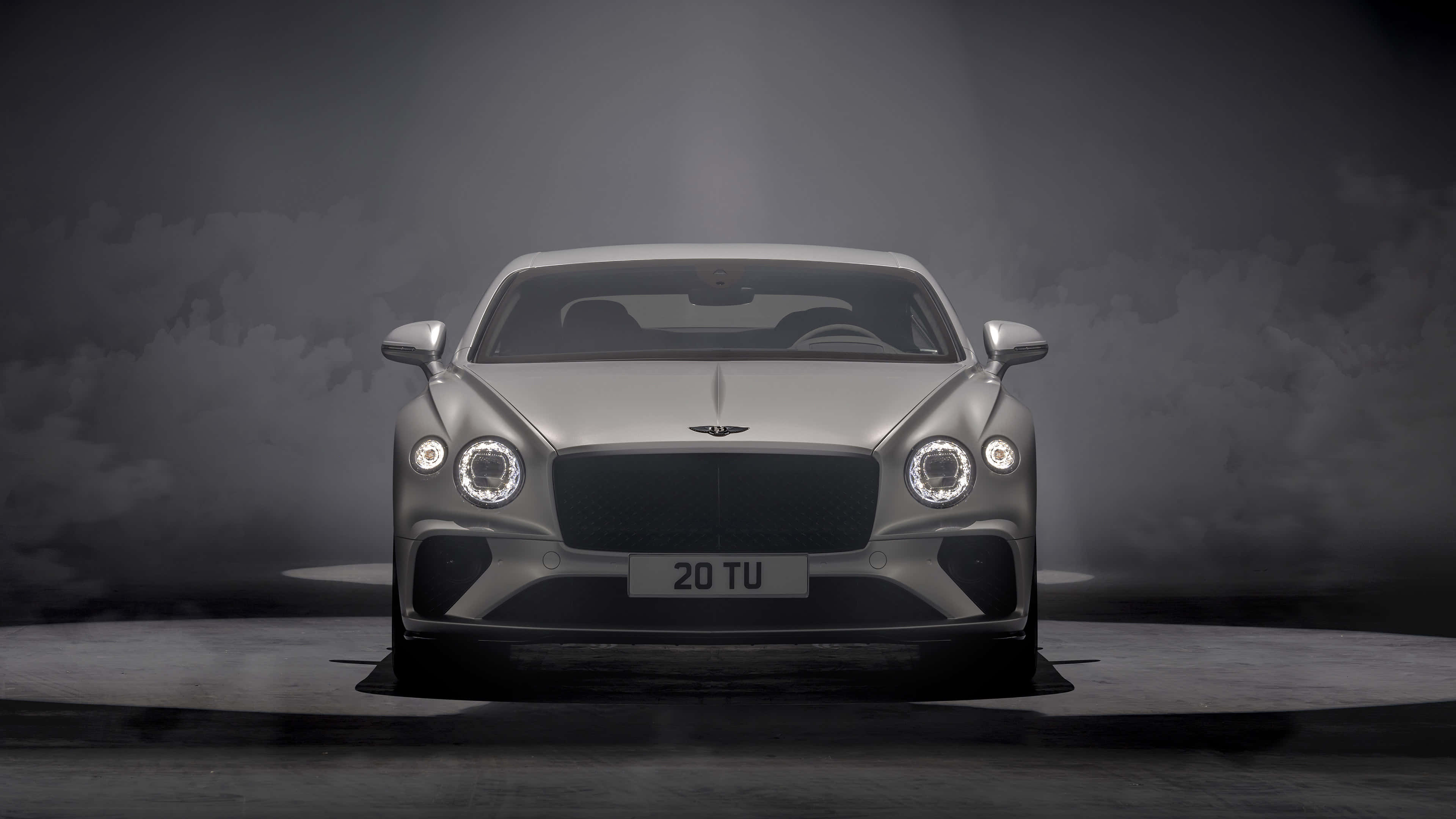 Bentley Continental Gt 桌布 壁紙 — 一輛時尚的銀色賓利 Continental GT 在戲劇性的聚光燈下，面向煙霧瀰漫的黑暗背景。