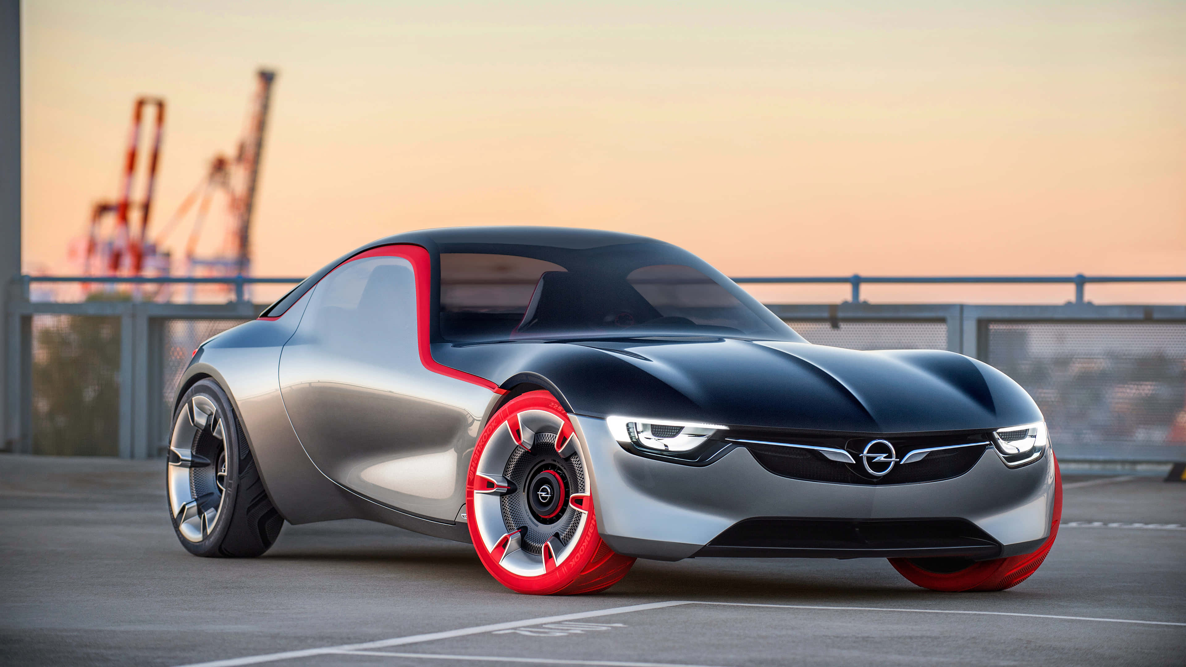 Opel Gt 桌布 壁紙 — 一輛時尚的銀黑色概念車，帶有紅色點綴，停在夕陽下的屋頂上，背景有起重機。